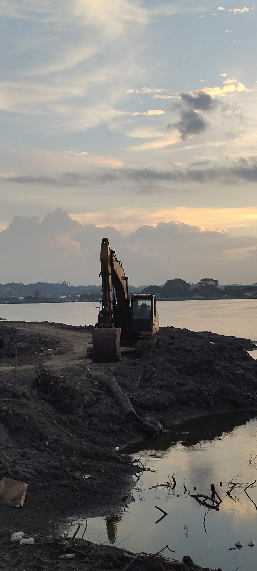 An excavator sits on a pile of dark, wet earth beside a body of water. The excavator is yellow and brown, with a large bucket at the end of its arm. The water reflects the sky, which is filled with dramatic clouds tinged with orange and yellow from the setting sun. In the background, across the water, is a distant shoreline with trees and some buildings. The foreground shows a muddy bank with debris, including a cardboard box and several plastic bottles. The scene appears to be a construction or excavation site near the coast.