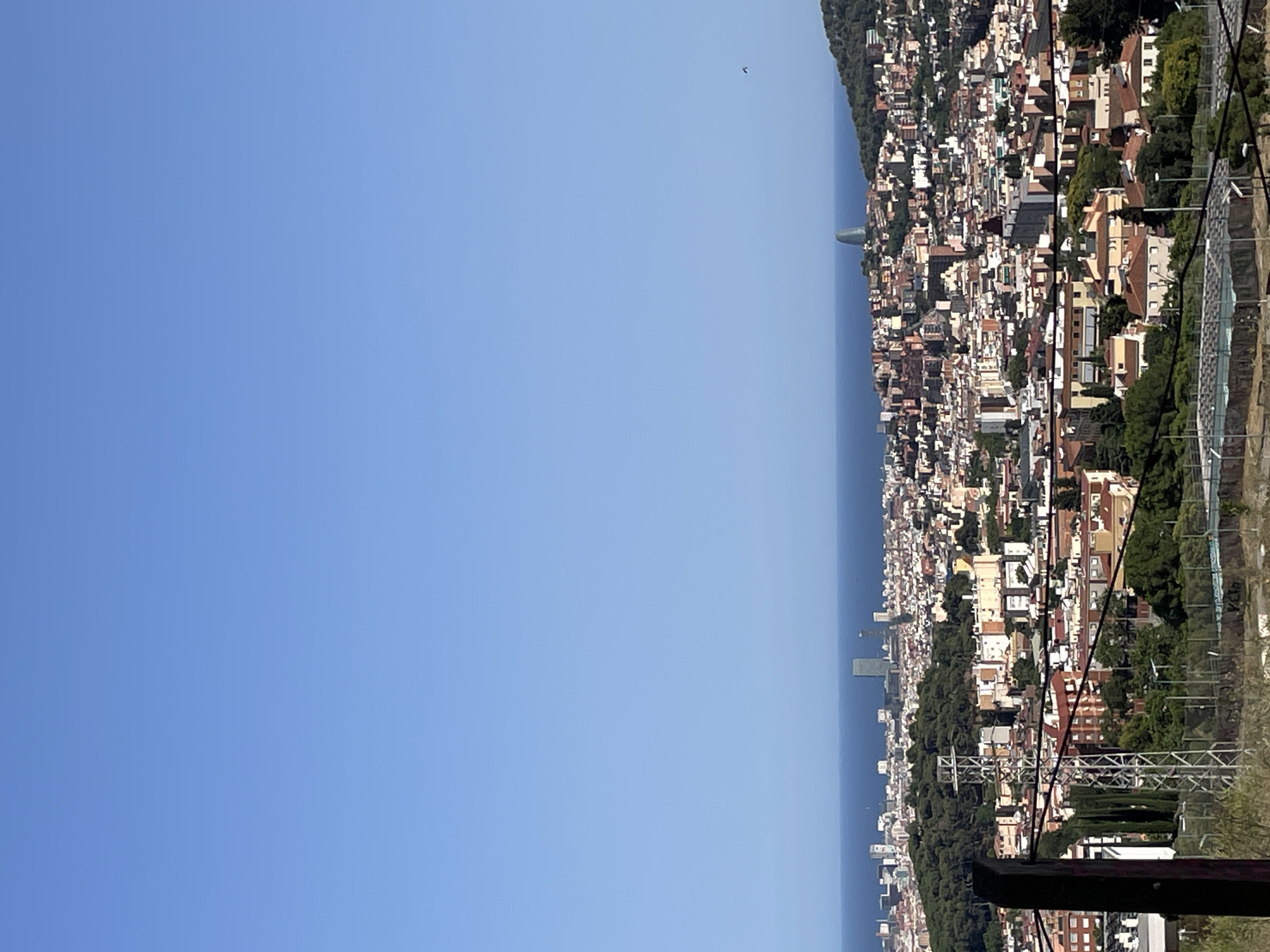 This is a high-angle, eye-level, wide shot of the cityscape of Barcelona, Spain, on a clear, sunny day. The sky is a vibrant, cloudless blue, taking up approximately two-thirds of the frame. Below the sky is the deep blue expanse of the Mediterranean Sea, meeting the horizon.
The city sprawls across the landscape, a dense collection of buildings with red-tiled roofs and white or light-colored facades. The architecture varies, with some buildings appearing older and more traditional, while others are modern high-rises. A distinctive, curved skyscraper, resembling a twisted bottle, stands prominently in the middle-right of the distant cityscape, a recognizable landmark of Barcelona. The city extends along the coastline and climbs up a green, tree-covered hill in the background.
In the foreground, a black wooden post and some black wires create a partial barrier, suggesting the vantage point is from a raised area or balcony, possibly a viewpoint or park. Green trees and some construction-like structures are visible closer to the viewer, adding depth to the scene. The overall impression is one of a bustling, sunny urban environment set against a beautiful coastal backdrop. There are no people visible in the image.
