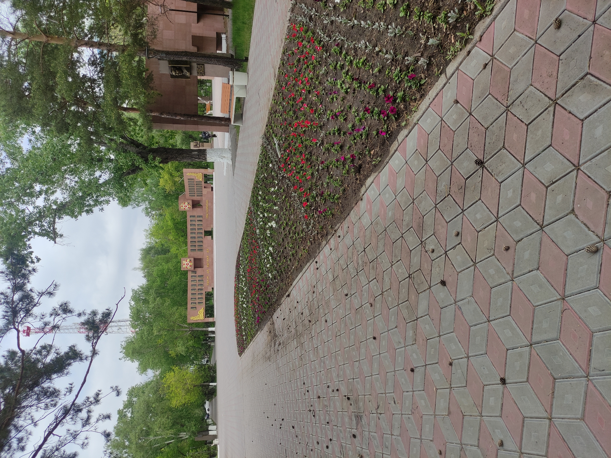 The image shows a pathway in a park or memorial complex in Pavlodar, Kazakhstan. The pathway is paved with alternating pink and gray geometric pattern blocks. A flowerbed borders the pathway, containing a variety of small flowering plants in shades of red, white, and purple. Scattered pine cones lie on the paved path. In the background, there are several commemorative structures, possibly war memorials, constructed from a dark red-brown stone or brick. Trees are visible on either side of the pathway. In the far background, a white structure or building can be seen. The sky appears overcast.
