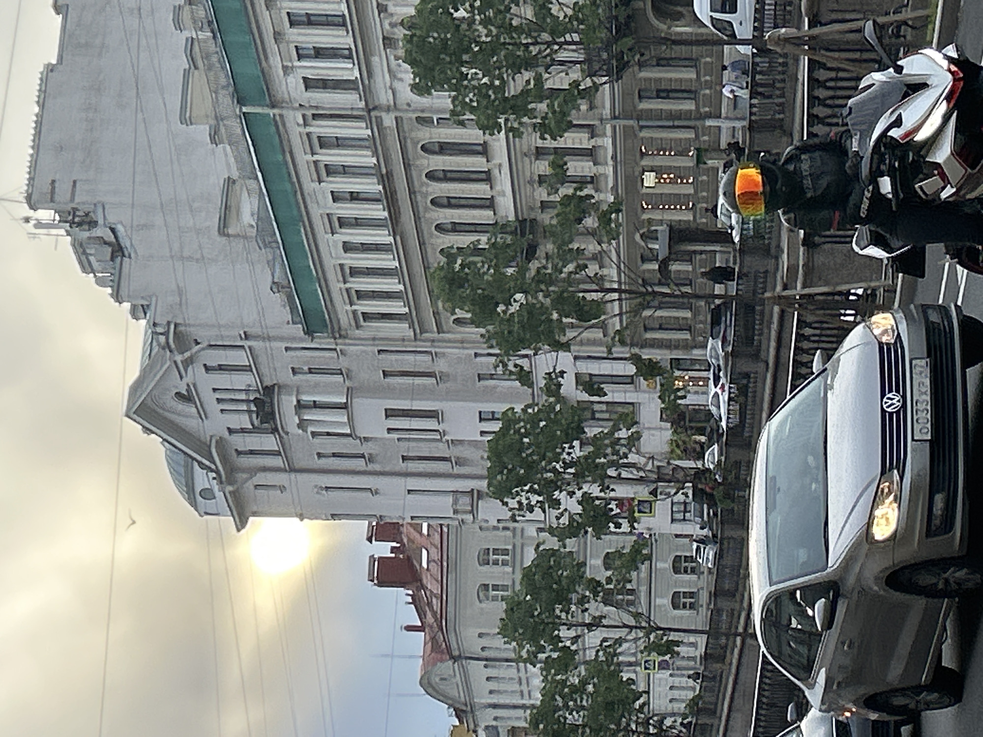 In an urban environment, a motorcyclist wearing a helmet with a reflective orange visor is stopped at what appears to be an intersection. The motorcyclist is dressed in dark motorcycle gear. In front of the motorcycle is a silver sedan, a Volkswagen, with its headlights on. The car has a license plate that reads "0033 XP 62". Behind the vehicles, there is a row of multi-story buildings with classical architectural elements, featuring arched windows and ornate facades. Trees line the sidewalk in front of the buildings, and a body of water, possibly a canal, is visible beyond the sidewalk with a decorative railing. The sky is overcast with a bright, diffused sun visible through the clouds. A bird is flying in the distance. There are other vehicles and pedestrians in the background, suggesting a busy city street.