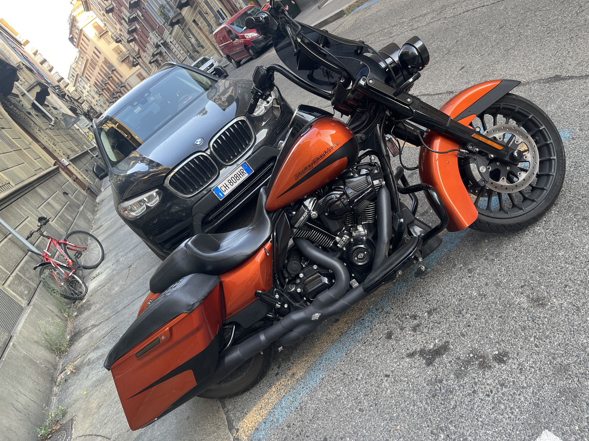 An orange and black Harley-Davidson motorcycle is parked on a city street in Turin, Italy. The motorcycle is angled towards the left of the frame. A black BMW SUV is parked behind the motorcycle, facing the same direction. The license plate of the BMW is visible and reads "CH 808HR". In the background, there are buildings typical of an Italian city and a red bicycle is leaning against a wall. The street is paved and shows some wear and tear, with blue and yellow paint markings on the ground. The lighting suggests it is daytime, possibly late afternoon due to the warm tones of the sunlight.