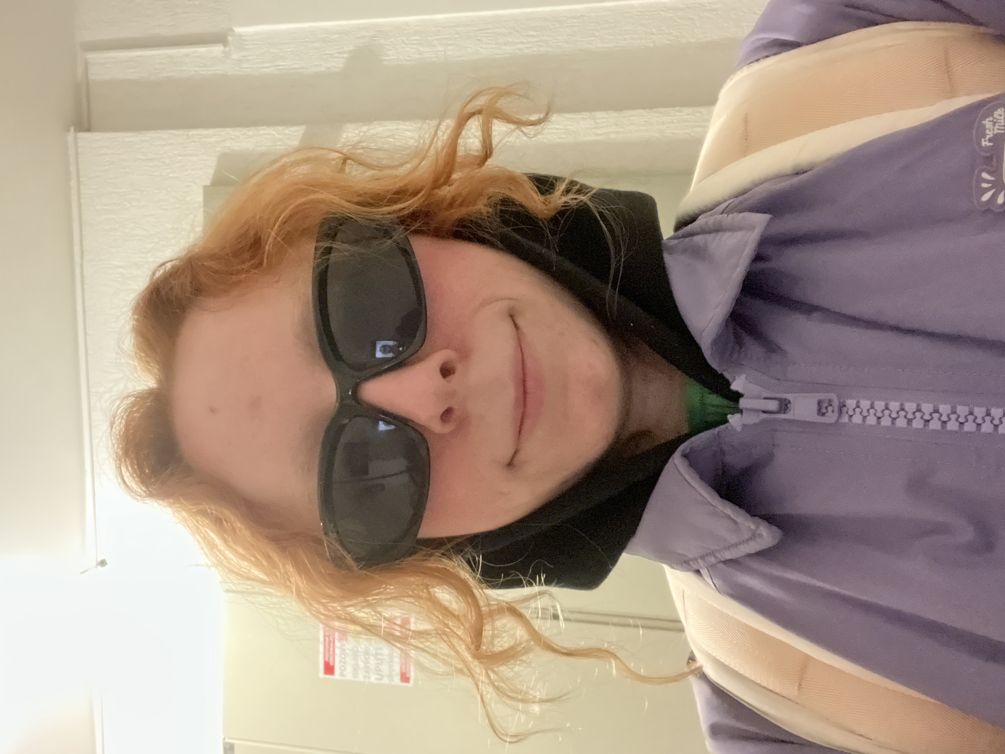 A person with wavy, reddish-blonde hair is wearing sunglasses, a black hooded sweatshirt, and a light purple jacket with a white zipper and collar. They appear to be indoors, with a white door and wall visible in the background. There's a sticker or label on the door with some red and white text. The location is unspecified, but based on the instructions, it is presumed to be somewhere in the city of Palilula Urban Municipality, Serbia.