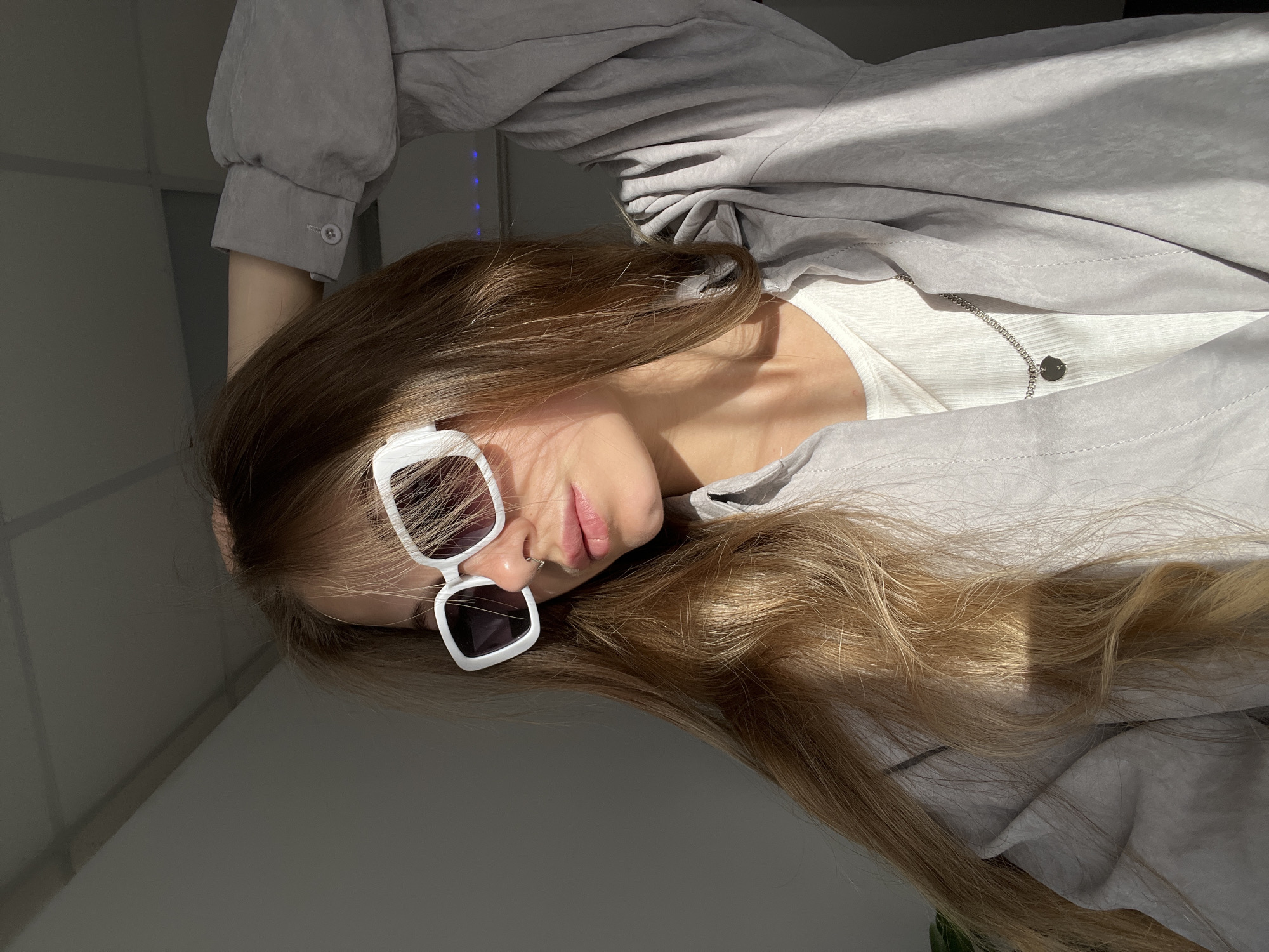 A person with long brown hair, wearing white-rimmed sunglasses, lies on a surface partially lit by sunlight. The person is wearing a white top and a light grey jacket. A silver necklace with a round pendant is visible. The background consists of a wall and ceiling. The location is not discernible.