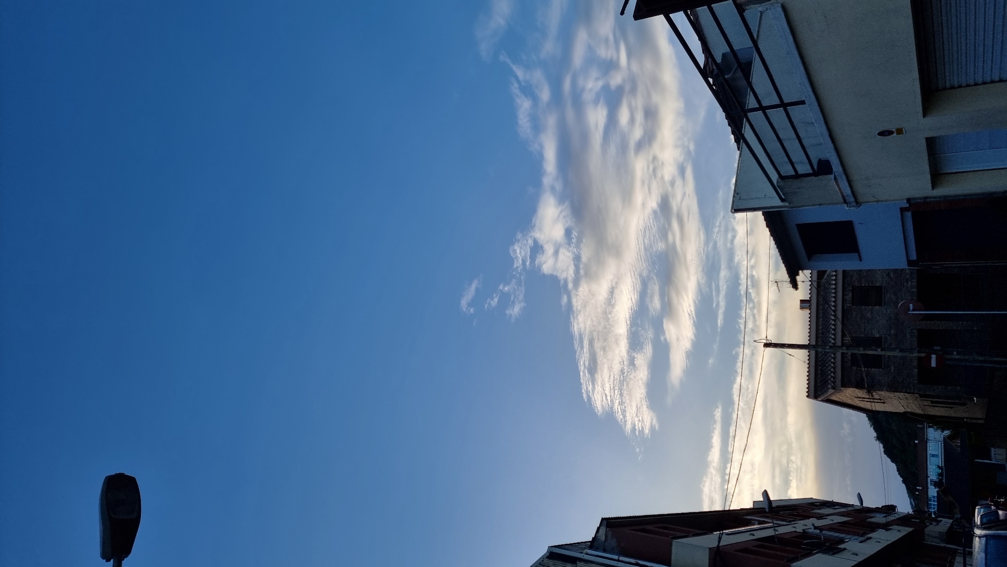 In Sant Joan Les Fonts, Spain, a vertical, low-angle shot captures a twilight sky with scattered clouds above a streetscape. A dark street lamp extends from the upper left corner. Buildings line the right and left sides of the frame, including a multi-story building on the far left with red and gray facades, and structures on the right with light yellow and light blue exteriors, one featuring a black metal railing. A dark brick building with visible windows is centrally located, partially obscured by foreground elements. Power lines stretch horizontally across the middle ground. The background features a distant, dark green hillside. The scene is devoid of human presence.