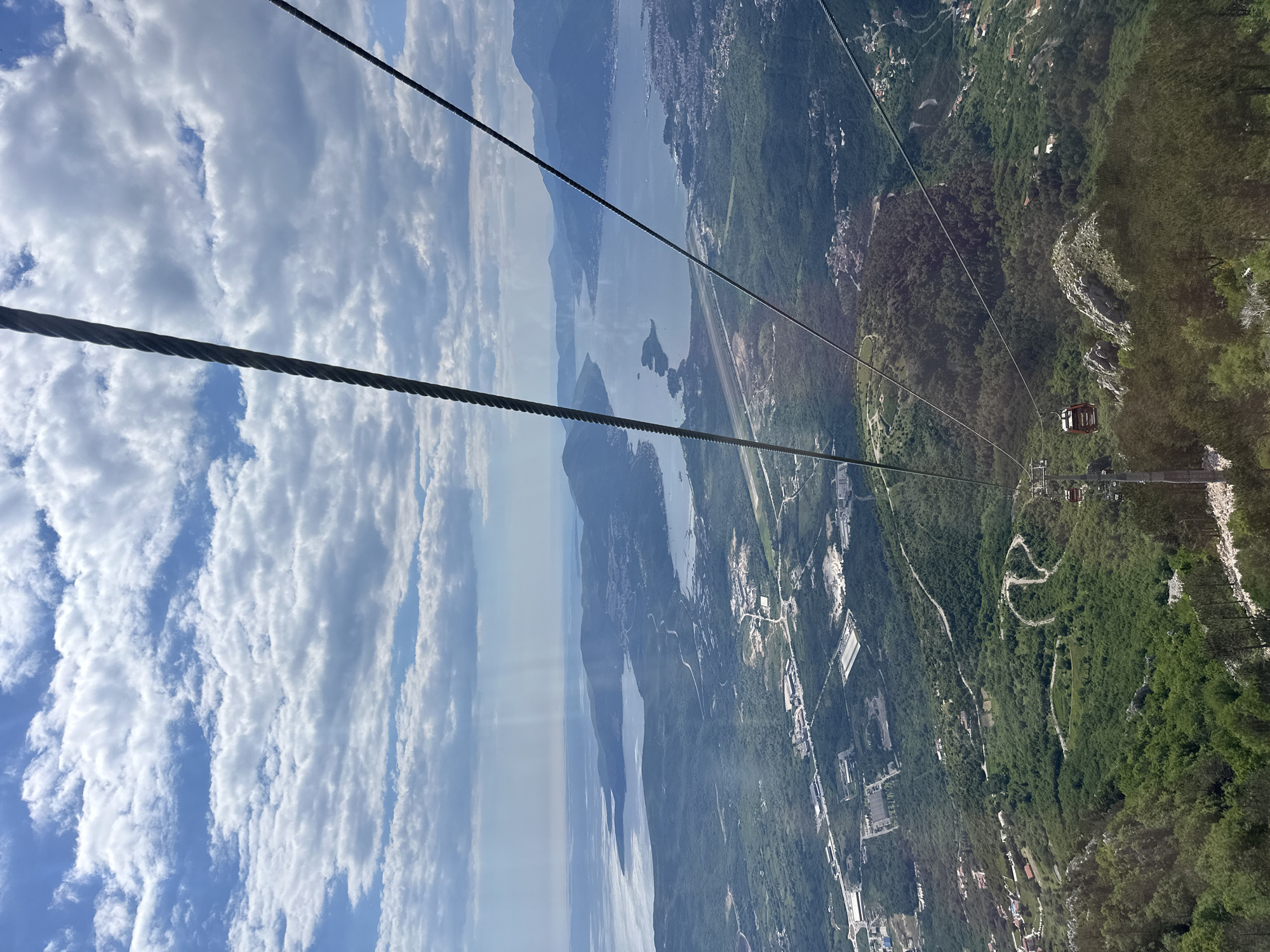 The image shows a view from a cable car ascending or descending in Škaljari, Montenegro. The scene features a panoramic vista encompassing the coastline, the Bay of Kotor, and mountainous terrain. Visible below are buildings, roads, and dense green vegetation. The sky is partly cloudy. The cable car wires stretch across the scene, leading to other cars.