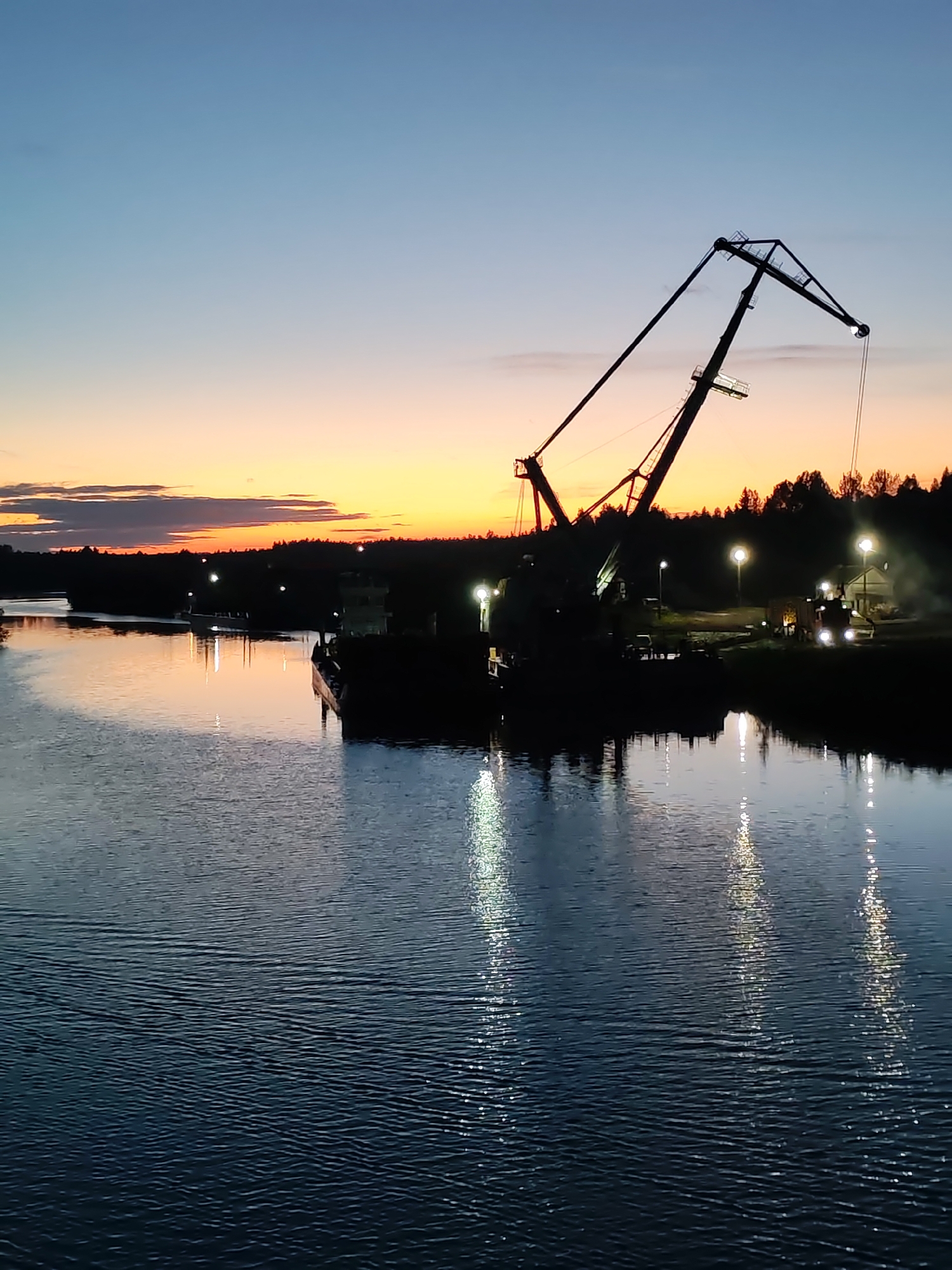 The image depicts a waterfront scene in Annensky Most, Russia, at twilight. A large, dark silhouette of a crane dominates the composition, positioned on a barge on the river. The sky transitions from dark blue to orange and yellow near the horizon, indicating either sunset or sunrise. Reflections of streetlights and the crane are visible in the river's surface. On the right side of the frame, there's a forested area with illuminated street lamps and vehicles. The water in the foreground exhibits ripples. A barge is visible in the background.