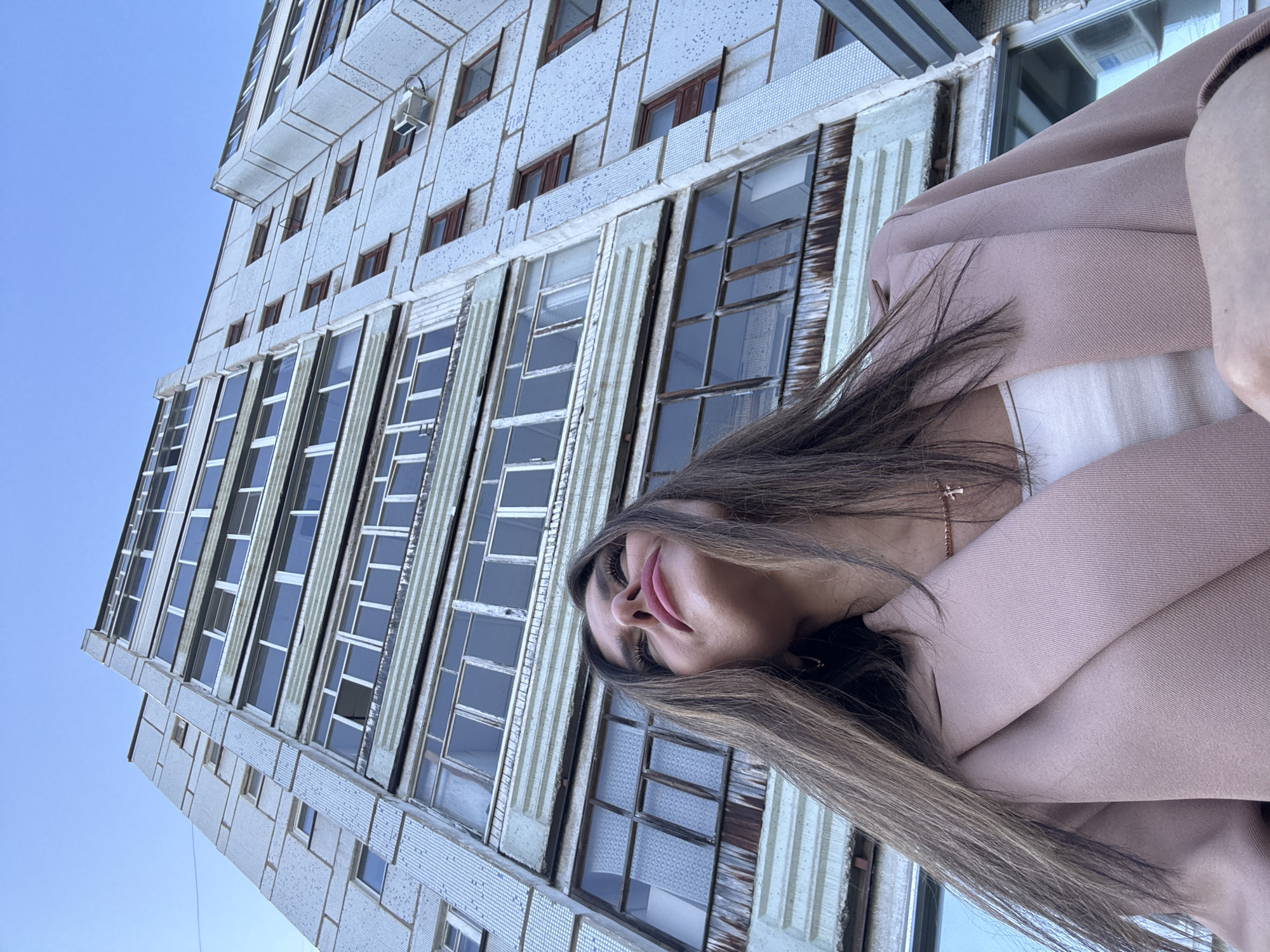 A low-angle shot captures a woman with long, light brown hair looking upwards against a backdrop of a multi-story residential building in Chaikovsky, Russia. The building, likely constructed with concrete and tile, features multiple balconies and windows, some of which appear weathered. The woman is wearing a light pink blazer over a white top and a delicate necklace with a cross pendant. The sky above is a clear blue. The perspective emphasizes the scale of the building and the upward gaze of the woman.