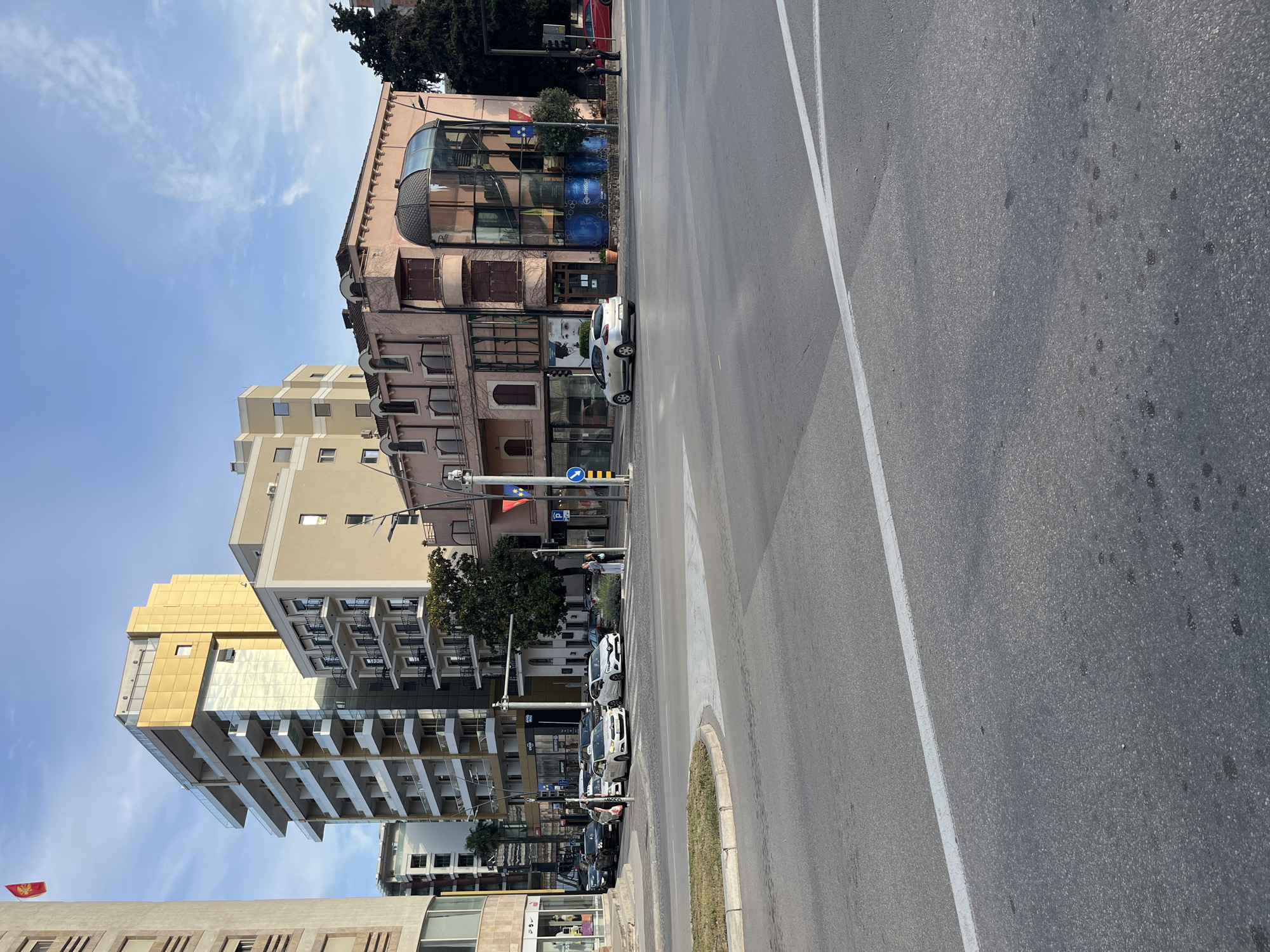 The image shows a street scene in Budva, Montenegro. On the left side of the road, there are several multi-story buildings, varying in color and architectural style. Some buildings are modern with glass balconies and golden facades, while others are older with pink facades and arched windows. Cars are parked along the sidewalk on the left. A street with white painted dividing lines runs along the right of the frame. The sky is blue with some clouds. There appear to be traffic lights, and there are pedestrians on the sidewalks.