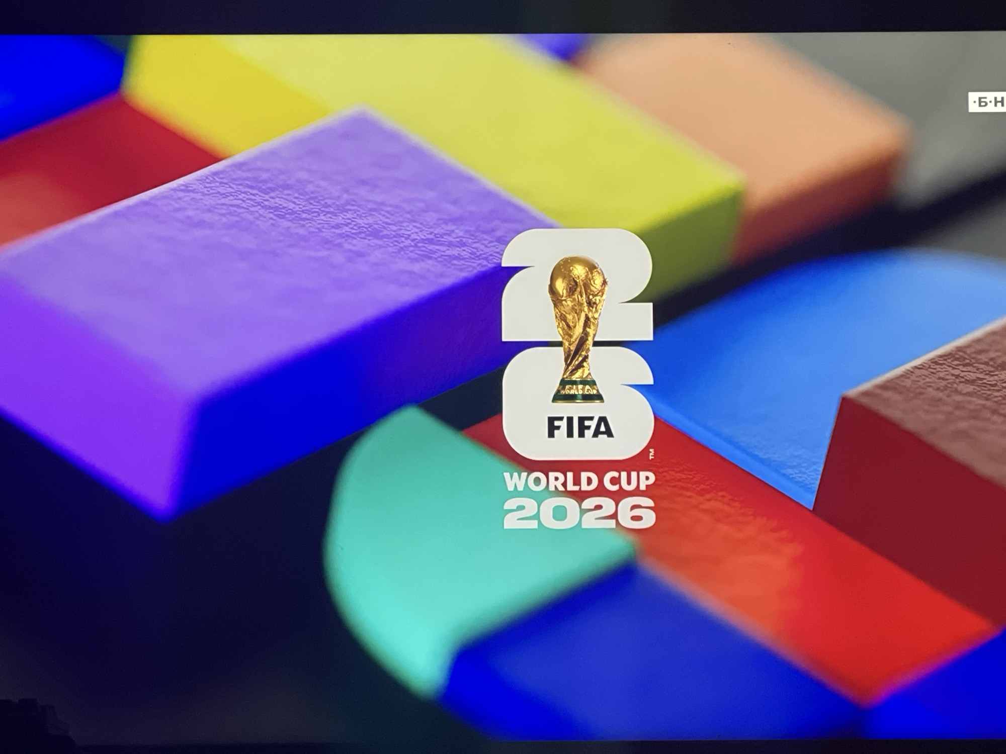 The image displays the logo for the FIFA World Cup 2026 superimposed on a background of colorful, overlapping geometric shapes. The logo features the FIFA World Cup trophy, the text "FIFA," and the numbers "26," all in white. Below this, "WORLD CUP 2026" is written in white text. The background consists of abstract, block-like shapes in shades of blue, yellow, purple, red, and orange, arranged in a dynamic, overlapping manner. The lighting suggests a digital display or a screen. There is no indication of the city of Ruse, Bulgaria, in this image.