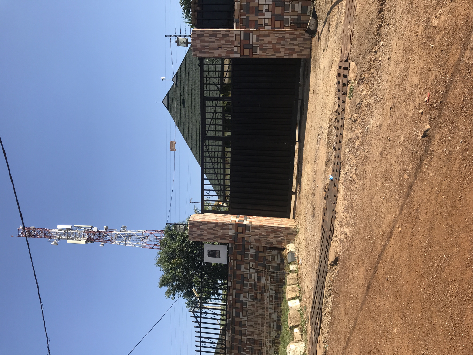 The image displays an outdoor scene featuring the entrance to a property, a dirt road, and a prominent telecommunications tower under a clear blue sky.
In the center, a dark metal gate serves as the entrance, flanked by sturdy gate posts decorated with a mosaic of multi-toned tiles in shades of brown, beige, and white. Behind the gate, a building with a dark green, pitched roof covered in tiles is partially visible. A black metal picket fence extends from the gate, with a verdant tree growing behind it to the left. An outdoor light fixture is mounted above the gate, and another is on the left gate post.
To the far left, a tall telecommunications tower dominates the skyline, characterized by its red and white lattice structure and numerous antennas and dishes. Utility lines crisscross the upper part of the frame, with a small, light-colored rectangular object (possibly cardboard) hanging from one.
The foreground consists of an unpaved, reddish-brown dirt or gravel road. A metal drainage channel or grate runs along the road's edge, adjacent to the property. On the right gate post, a white box bears a black and white sign that clearly reads "NOTICE" at the top, with additional text below that appears to be "PROPERTY" or "PREMISES."
The scene is well-lit by bright sunlight, indicating daytime with clear weather. No people or active events are present, conveying a static view of a residential or commercial area in what appears to be a developing region. The location context is Mandate Estate, Ilorin, Nigeria.