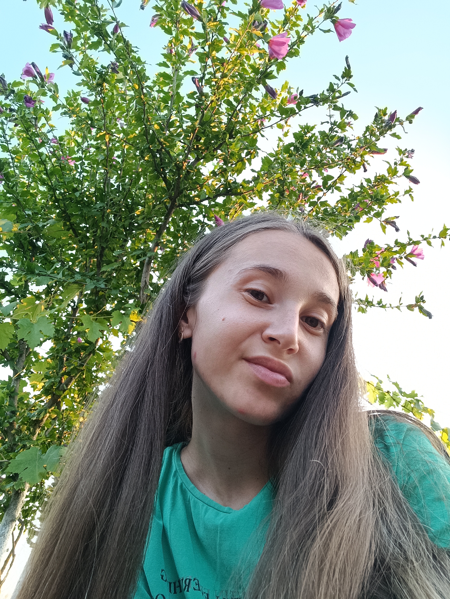 A young woman with long, brown hair is pictured in a close-up shot outdoors, with a tree and flowers in the background. She is wearing a green t-shirt. The location is Vypasne, Ukraine.