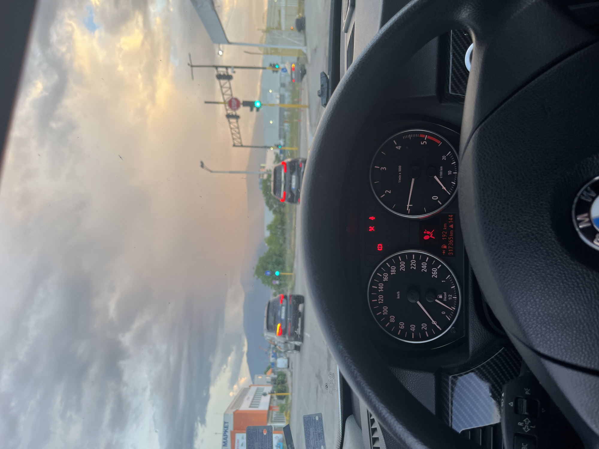 The image is a vertical, wide-angle shot taken from inside a car, looking forward through the windshield and at the dashboard. The primary subjects are the car's interior and the road scene outside.
In the foreground, a portion of the steering wheel is visible on the right, with a BMW logo on the center. The dashboard includes a speedometer and a tachometer. The speedometer is marked in km/h, showing a needle pointing between 180 and 200 km/h. The tachometer is marked in 1/min x 1000, with the needle between 2 and 3. Text on the dashboard indicates "Diesel", "192 km", and "317365km". Red warning lights are visible on the dashboard.
Outside the car, the scene depicts a road with traffic lights, cars, and buildings. A traffic light with a green signal is visible. Several cars are on the road, with one black car directly ahead. The sky is filled with dramatic clouds, suggesting either sunrise or sunset, with patches of yellow and grey. In the distance, trees and mountains are visible. On the left side of the road, a building with an orange and white facade is labeled with the text "MAPKET".
The setting appears to be a road intersection in an urban or suburban area, possibly on the outskirts of Sofia, Bulgaria, given the location context. The time of day is likely early morning or late evening due to the lighting conditions and the appearance of the sky. The overall mood is somewhat neutral, focusing on the driving experience and the surrounding environment.
