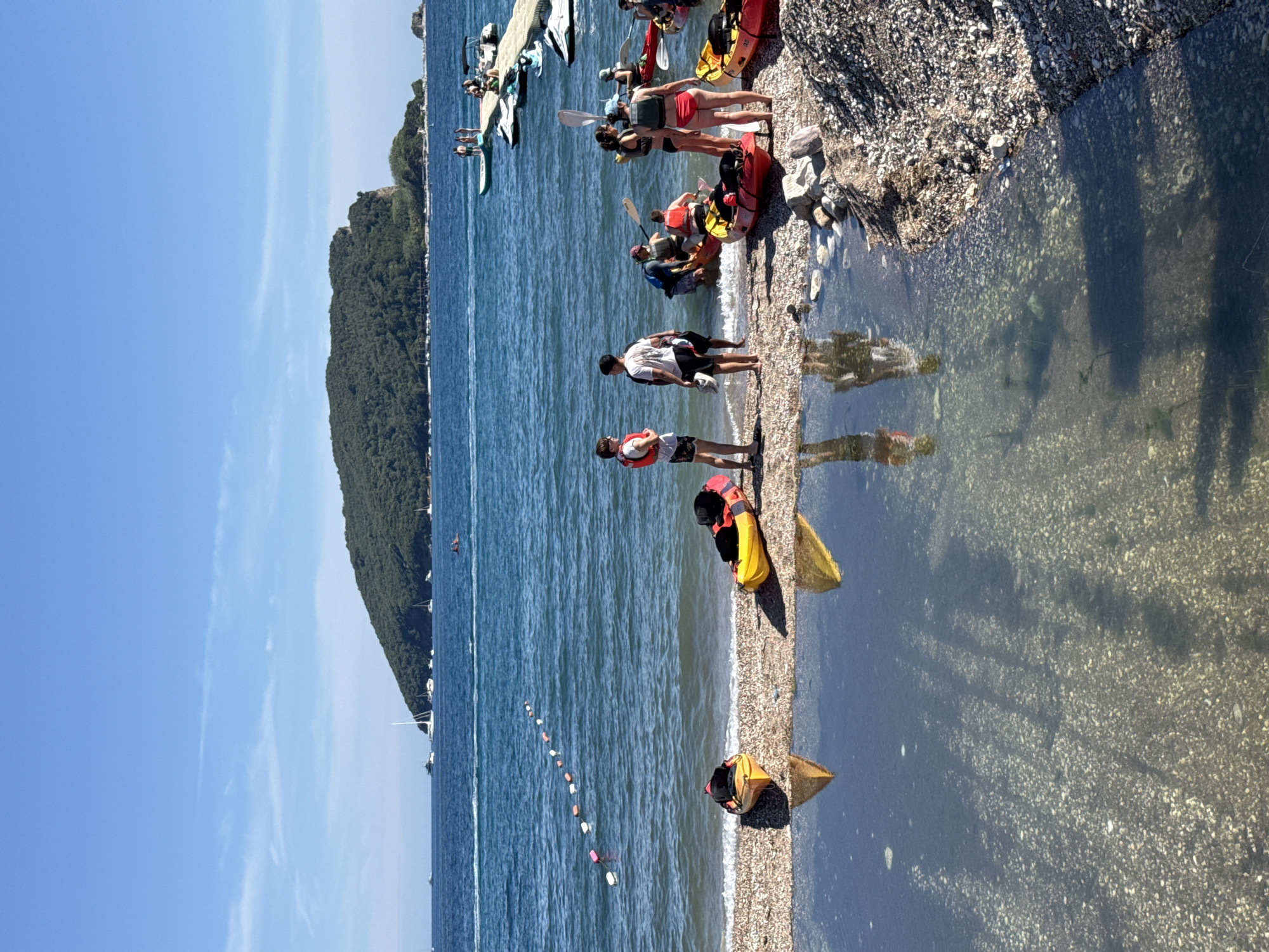 The image depicts a sunny day at a beach in Budva, Montenegro, with a group of people preparing to go kayaking. In the foreground, two kayaks are pulled up onto the pebble beach. The water is clear, and the reflections of the kayaks and people are visible on its surface. Several individuals are gathered near the water's edge, some in kayaks and others standing on the shore, dressed in swimwear and life vests. A floating rope with buoys separates the shallow water from the deeper sea. In the background, a large, tree-covered island dominates the horizon, and several sailboats and a modern pier with jet skis are visible in the distance. The overall scene conveys a sense of leisure and outdoor activity.