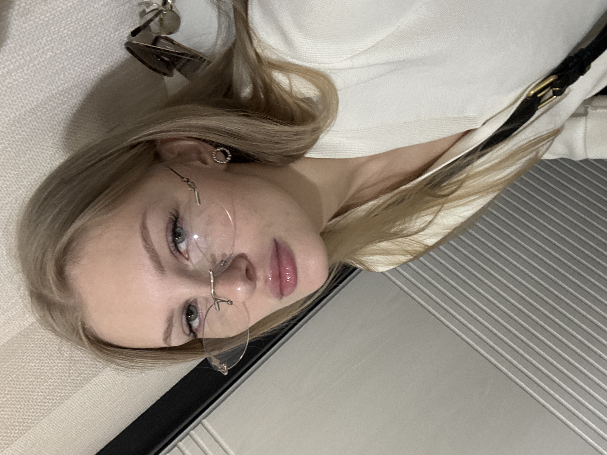 A young woman with blonde hair and blue eyes, wearing glasses and a white top, poses for a selfie. She has on subtle makeup, including lipstick and eyeliner. A circular, textured earring is visible on her left ear. She is positioned in front of a textured wall with vertical lines, possibly in an indoor setting in Perm, Russia.