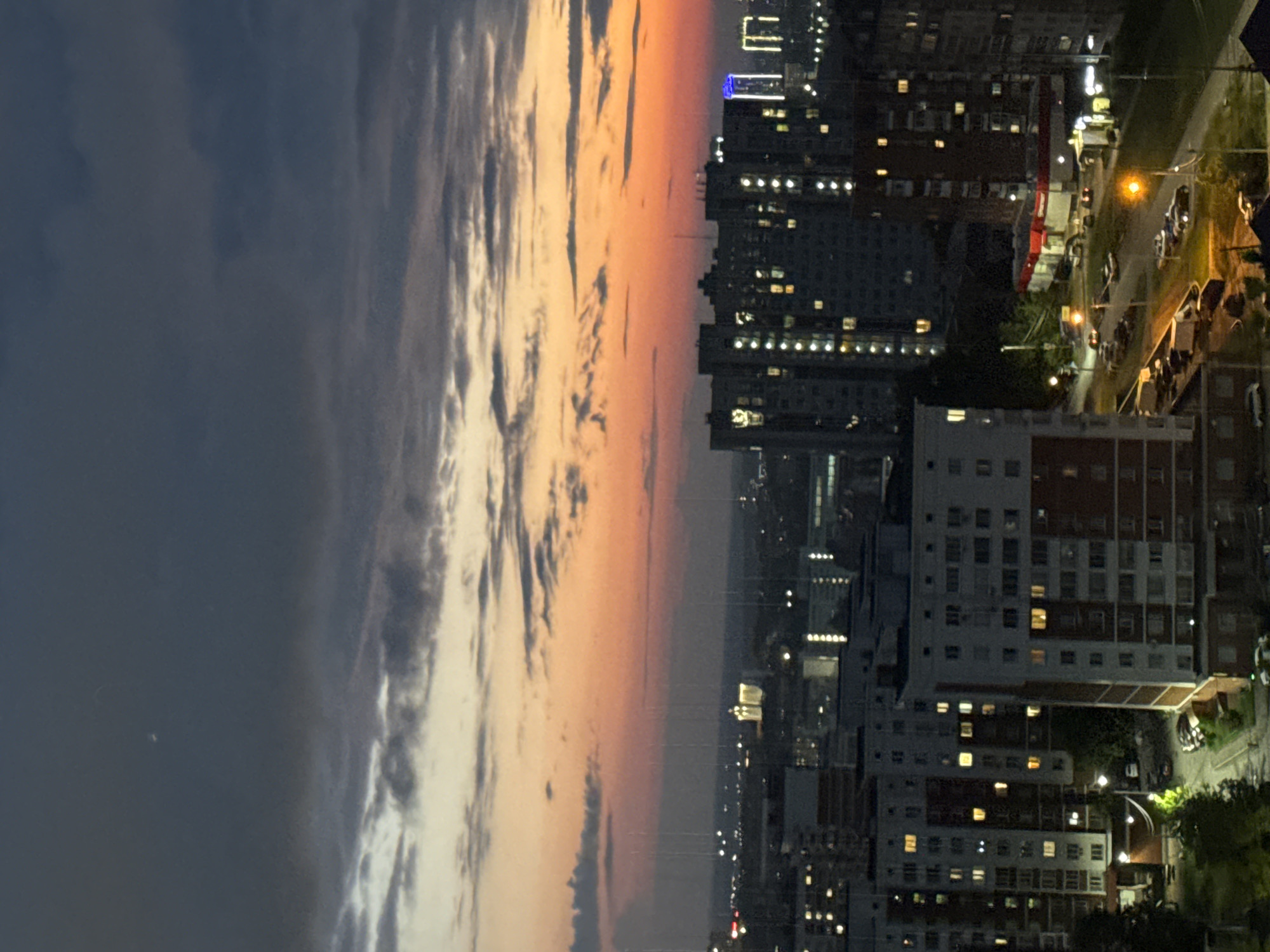 The image depicts an aerial view of a residential area in Izhevsk, Russia, during twilight. The sky is filled with dark, dramatic clouds, transitioning to a vibrant orange and yellow sunset horizon. Several multi-story apartment buildings dominate the cityscape, with numerous windows illuminated, indicating occupancy. In the distance, a brighter, lighter colored building with a distinct architectural style stands out, possibly a landmark or a public facility. The foreground shows more apartment blocks, a street with parked cars, and scattered trees. The overall impression is that of a densely populated urban environment at the end of the day.