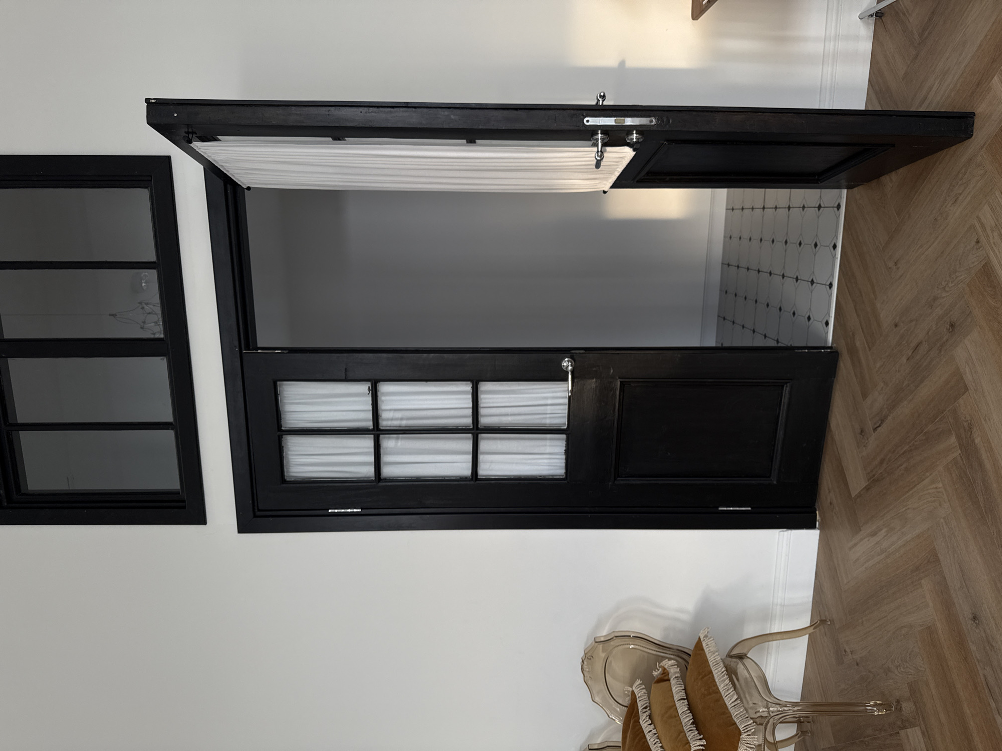 The image shows a partially open black door with multiple glass panes, each covered with white pleated blinds. The door is in the process of being opened, revealing a glimpse into a room with patterned floor tiles and white walls. To the left of the door, there is a large black-framed window with multiple panes. The scene appears to be indoors, possibly in a modern home or a commercial space. The flooring in the foreground is a light-colored wood laid in a herringbone pattern. Two transparent chairs with cushions are visible in the lower right corner of the image, partially obscured. The lighting suggests it might be daytime, with light casting shadows from the door frame onto the wall. No people are visible in the image. There is no visible text. The context provided indicates the location is Izhevsk, Russia.