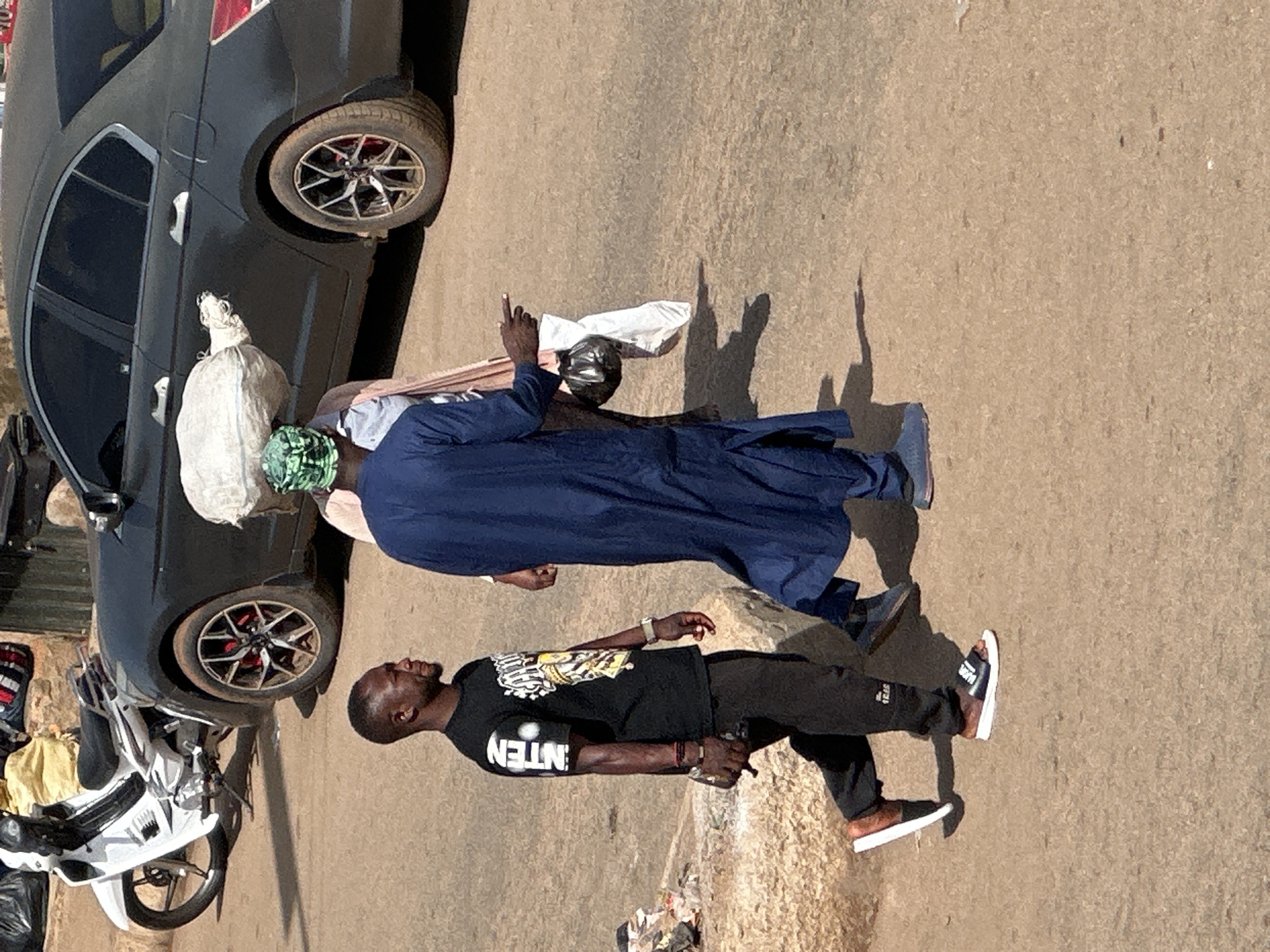 In Bukuru, Nigeria, two men are walking down a dusty street. The man on the left wears a black t-shirt with white lettering and black pants, while the man on the right is dressed in a blue robe. Between them, another person, partially obscured, carries a large white bag on their head. A dark sedan is parked nearby, and a white motorcycle is visible in the background. The ground is a dirt road, and there are some scattered debris and rocks. The scene captures a moment of everyday life and movement in an urban setting.