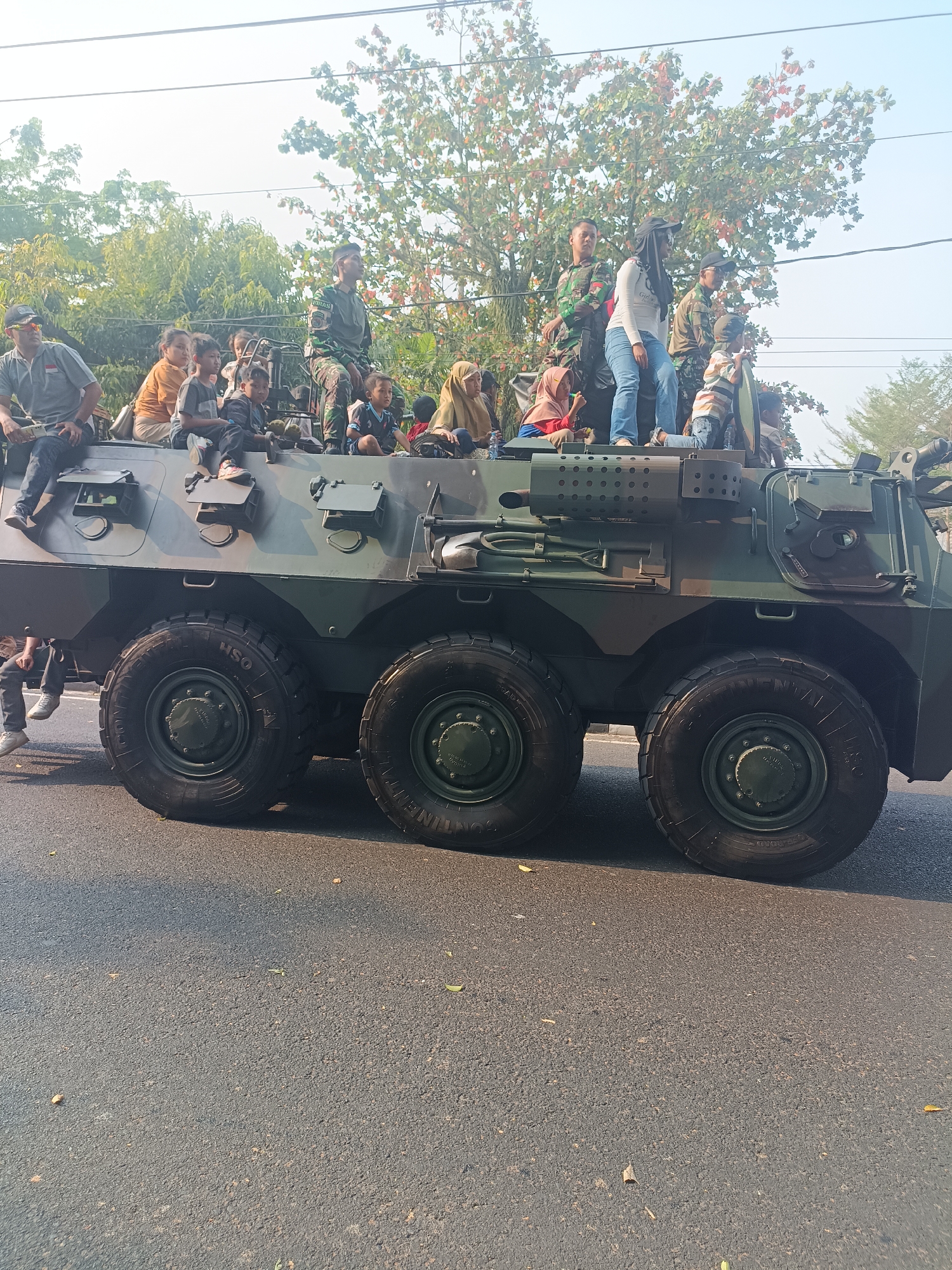 In Sleman, Indonesia, on a bright, sunny day, a camouflage military armored personnel carrier is parked on an asphalt road. The vehicle has six large, dark tires, with the front three visible on the right side of the image. Several people, a mix of adults and children, are sitting and standing on top of the APC. Among them are individuals in military uniforms and civilians in casual clothing, including women wearing hijabs. The scene appears to be a public event or parade, as indicated by the presence of civilians on the military vehicle. Trees with green and reddish-brown leaves line the background, and power lines are visible against the light sky. The overall impression is one of a community gathering or celebration involving the military.