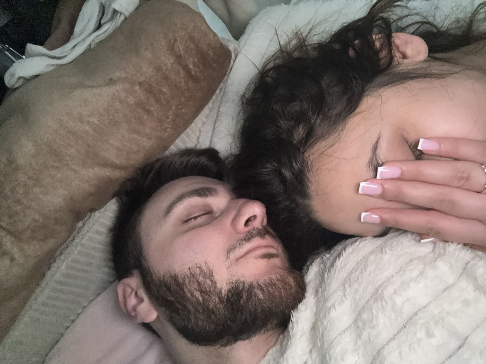 The image displays two individuals, a man and a woman, resting closely together indoors. The man, positioned in the foreground on the left, lies on his back with his eyes closed, suggesting he is asleep or deeply relaxed. He has a dark beard and dark hair. Next to him, on the right, the woman is also lying down, largely obscured by her dark, curly hair and her hand. Her left hand, adorned with French-tipped fingernails and a ring on her ring finger, is gently placed over her closed eyes. Both subjects appear peaceful and comfortable.
A fluffy, light-colored blanket or robe covers parts of both individuals. To the far left, a textured, possibly brown or beige pillow or blanket is visible, further indicating a bedding or resting environment. The lighting is soft and subdued, suggesting a low-light setting, likely night or early morning. The scene conveys a sense of intimacy and tranquility. There is no visible text in the image. The location context is Mönchengladbach, Germany.