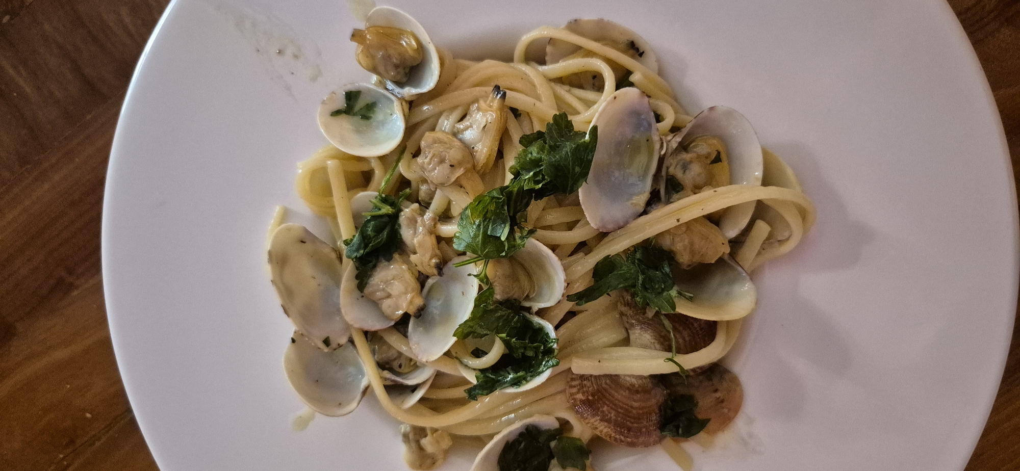 This is a top-down, close-up shot of a plate of linguine alle vongole (linguine with clams) situated on a white plate. The pasta is intertwined with numerous small clams, many of which are open, revealing their light-colored flesh. Scattered throughout the dish are chopped green herbs, likely parsley, which add a touch of freshness. The pasta appears to be coated in a light sauce. The background shows a portion of a wooden table, suggesting the dish is served in a restaurant or home setting. There are no people visible in the image. The dish is presented in a rustic yet appetizing manner, typical of Italian seafood cuisine. There is no information in the image to suggest it is located in the city of Taranto, Italy.