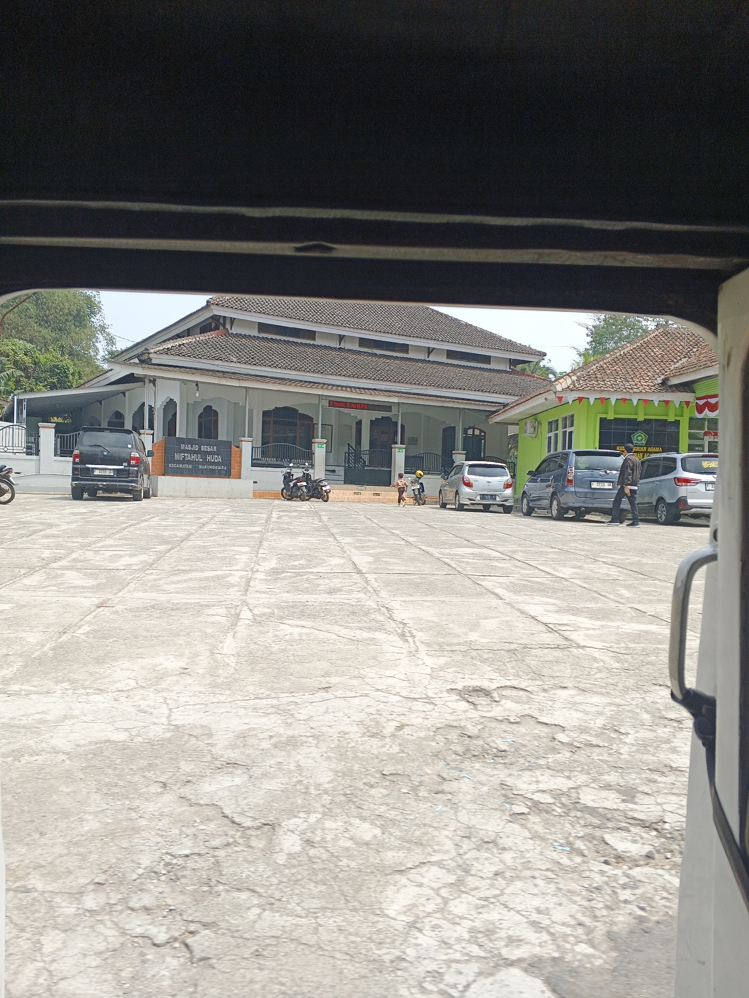 A view from inside a vehicle, looking out at a mosque and adjacent building in Warungkiara, Indonesia. The mosque, labeled "MASJID BESAR MIFTAHUL HUDA KECAMATAN WARUNGKIARA," is white with a brown tiled roof and appears to be the main structure. To its right is a bright green building with a sign partially visible that reads "KE...ERIAAN AGAMA". Several cars and motorcycles are parked in the paved courtyard in front of the buildings. A man in a dark jacket and pants is walking away from a blue minivan, and two children are visible playing near a motorcycle in front of the mosque. The ground is cracked concrete, and trees are visible in the background. The sky is overcast.