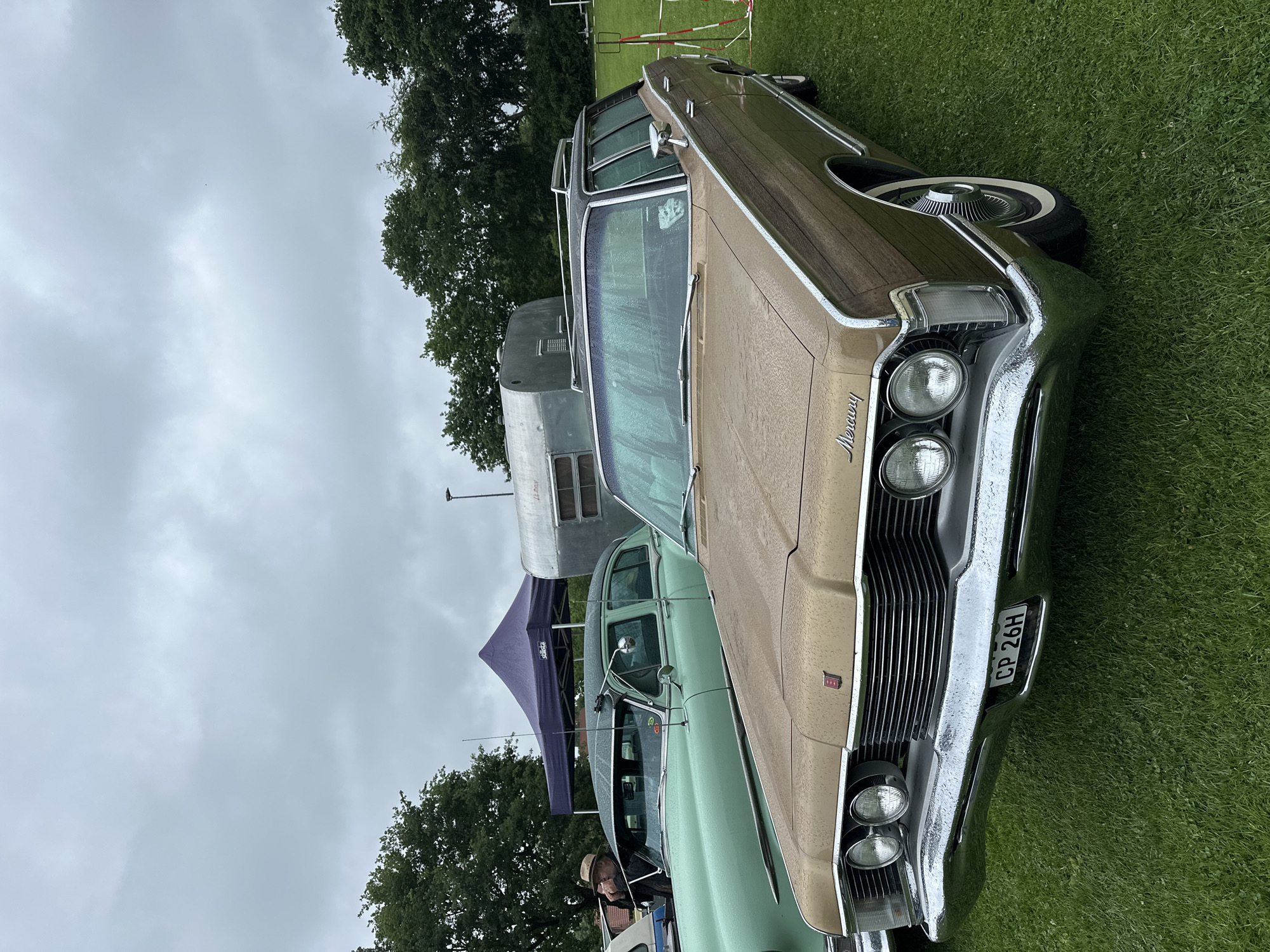 This image captures an outdoor gathering of classic cars on a cloudy, wet day in Deinste, Germany. The foreground is dominated by a brown 1960s Mercury station wagon, likely a Colony Park or Marquis, featuring faux wood paneling on its sides and a chrome front bumper. The car's hood, roof, and windshield are covered in numerous raindrops. The front grille is horizontal with quad circular headlights, and the "Mercury" script is visible on the left side of the hood. A white-on-black license plate, "CP 26H," is mounted below the grille. The car also has a chrome roof rack and whitewall tires.
To the left and slightly behind the Mercury, a light green, older classic sedan is visible, also wet with raindrops. A man wearing a hat is partially seen next to this green car, holding a small object to his mouth. Two whip antennas extend from the front of the green sedan.
In the mid-background, a purple pop-up canopy tent stands to the left of a vintage silver travel trailer with horizontal ribbing. Dark green deciduous trees line the horizon in the background, with an overcast, light grey sky above them. The ground is covered in green grass, which appears wet. A light pole and part of a white soccer goal are faintly visible in the far background on the right.