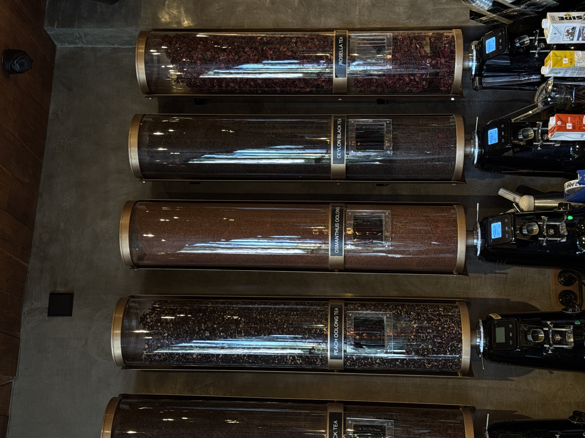 This image depicts a selection of teas in cylindrical dispensers, likely taken in a cafe or specialty store in the Ubud District of Indonesia. The dispensers are arranged vertically on a grey wall. Starting from the top, the labels indicate "ROSELLA TEA," "CEYLON BLACK TEA," "OSMANTHUS OOLONG," and "PEACH OOLONG TEA." Next to each dispenser, there's a dispensing machine with a digital display. The overall setting suggests a focus on high-quality tea offerings and a modern, minimalist aesthetic.