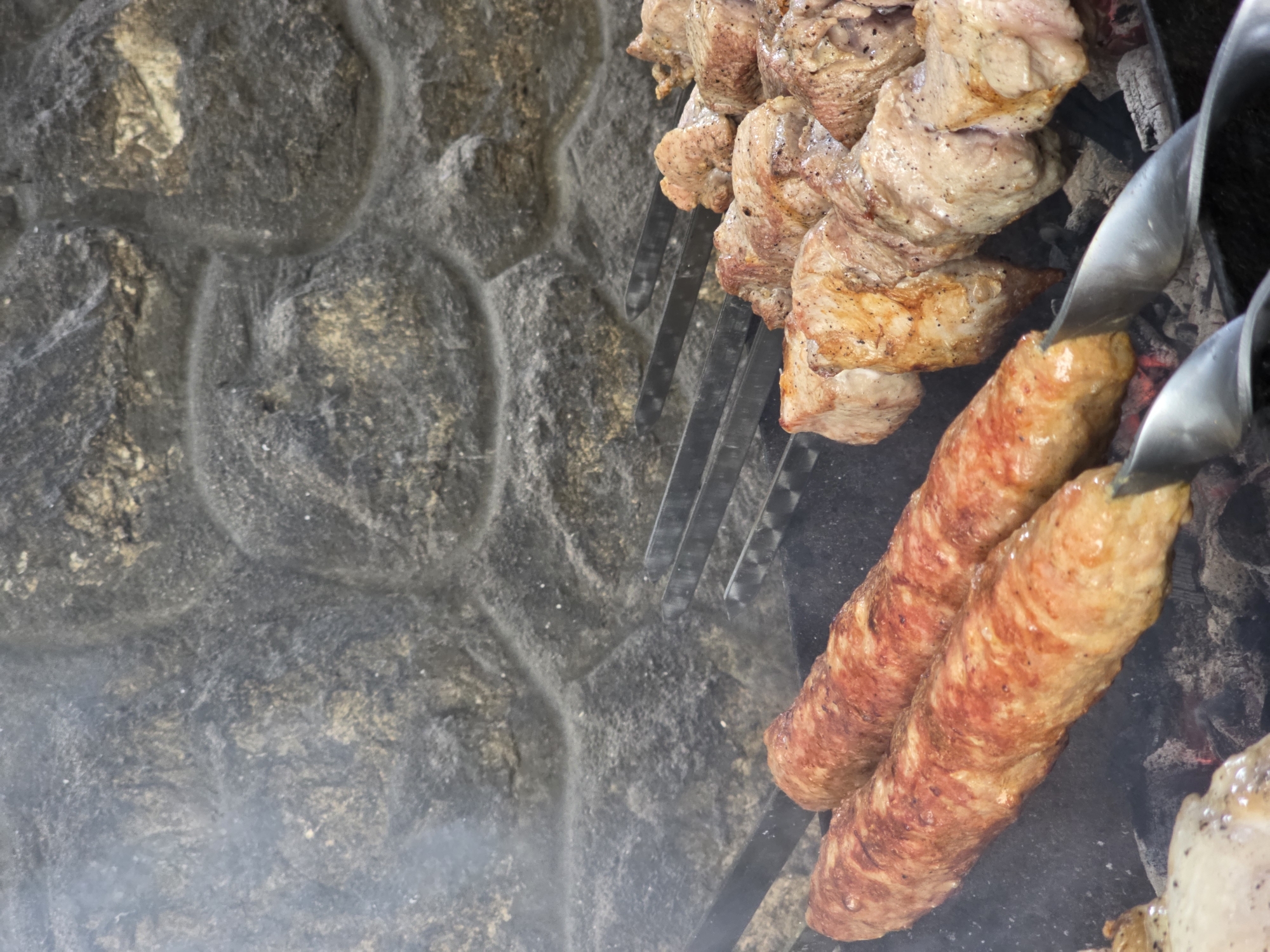 This close-up image shows several skewers of meat cooking over hot coals. The meat on the skewers appears to be a mix of cubed pieces of what looks like pork or lamb, and long, cylindrical sausages. The meat is browned and slightly charred in places, indicating it is in the process of being grilled. Smoke is visible rising from the coals, adding to the atmosphere of an outdoor barbecue.
The background of the image features a rough, textured stone wall, likely part of an outdoor cooking area or fireplace. The stones are varied in color and shape, with visible mortar lines. The overall impression is of a rustic, perhaps traditional, setting for preparing food.
The focus is on the food and the cooking process. There are no people visible in the frame, nor any clear indications of the time of day or specific weather conditions. The lighting suggests it is daytime, and the smoke implies an active cooking session. The location context provided places this scene in Pidhorodne, Ukraine, suggesting a possible cultural or culinary tradition of grilling meat. The image highlights the appetizing appearance of the grilling meat, with the focus on its texture and color. There is no visible text in the image.