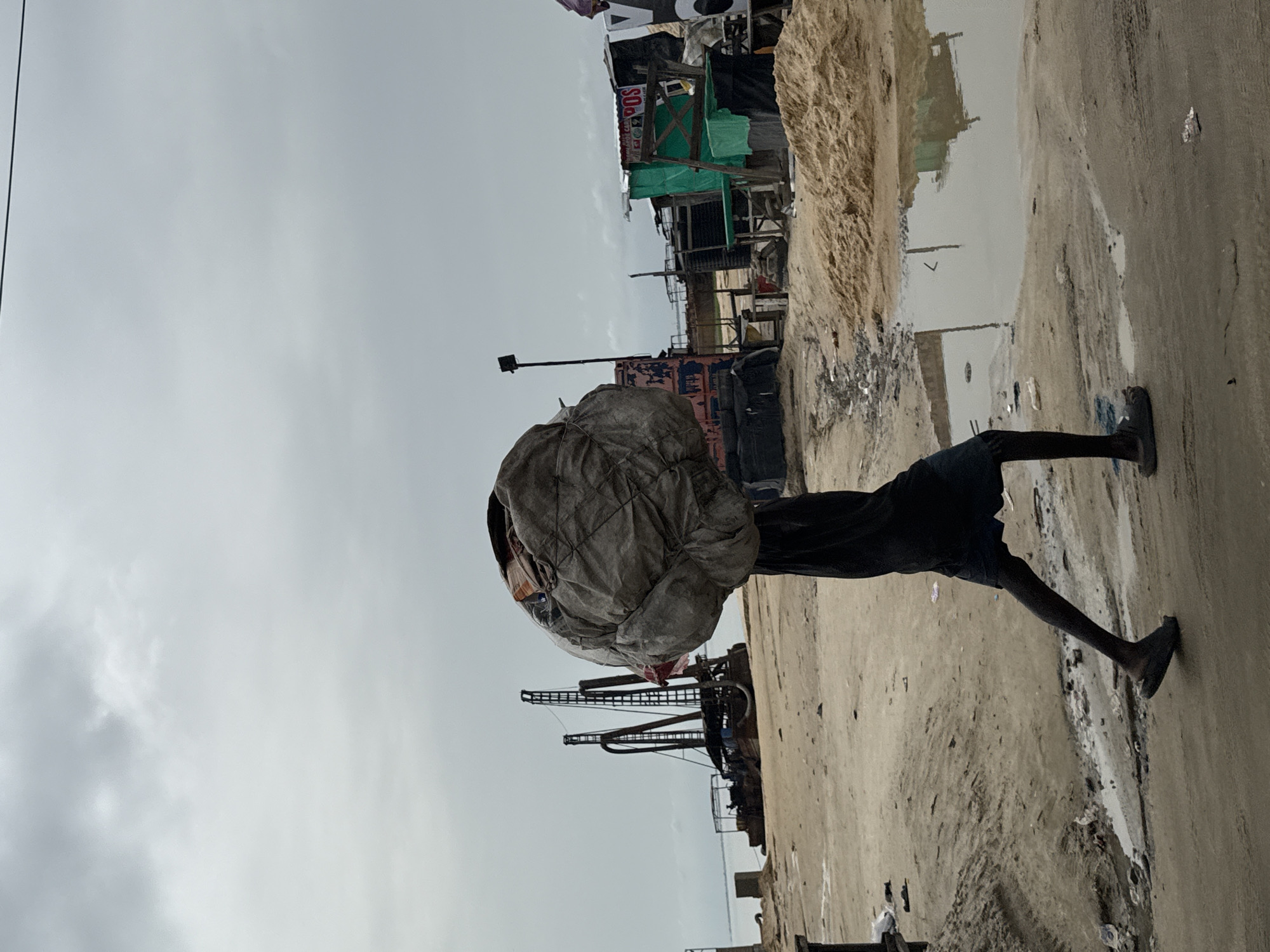 A person is walking along a street with a large, tied-up bundle on their head. The street has a wet, sandy surface with a large puddle to the right. Buildings and structures are visible behind the person, with a cloudy sky overhead. The scene appears to be in a city environment, identified as Ado, Nigeria.