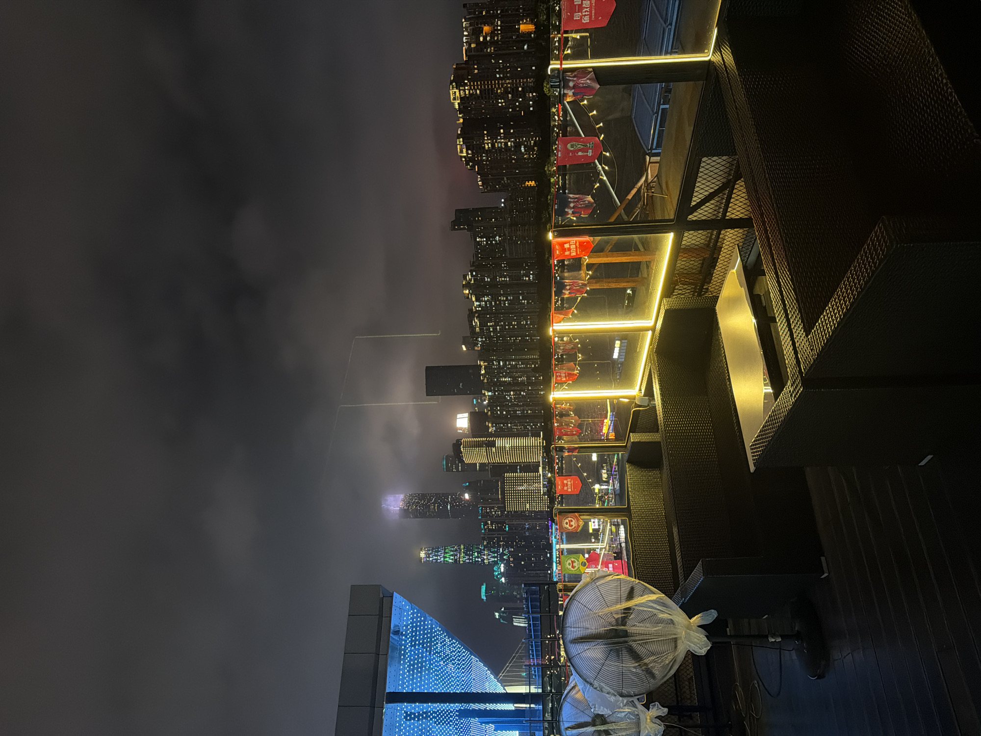 The image depicts a night view from a rooftop seating area in Haizhu District, China. The foreground features black rattan furniture, including couches and tables, illuminated by warm yellow strip lights along the balcony railing. Two large industrial fans are visible on the left, covered with plastic. In the background, a cityscape is illuminated by numerous lights from tall buildings, indicating a dense urban environment. A prominent bridge structure is visible on the far left. Several red banners with Chinese characters and images are hung along the railing. The sky is dark and cloudy, with some light pollution from the city.