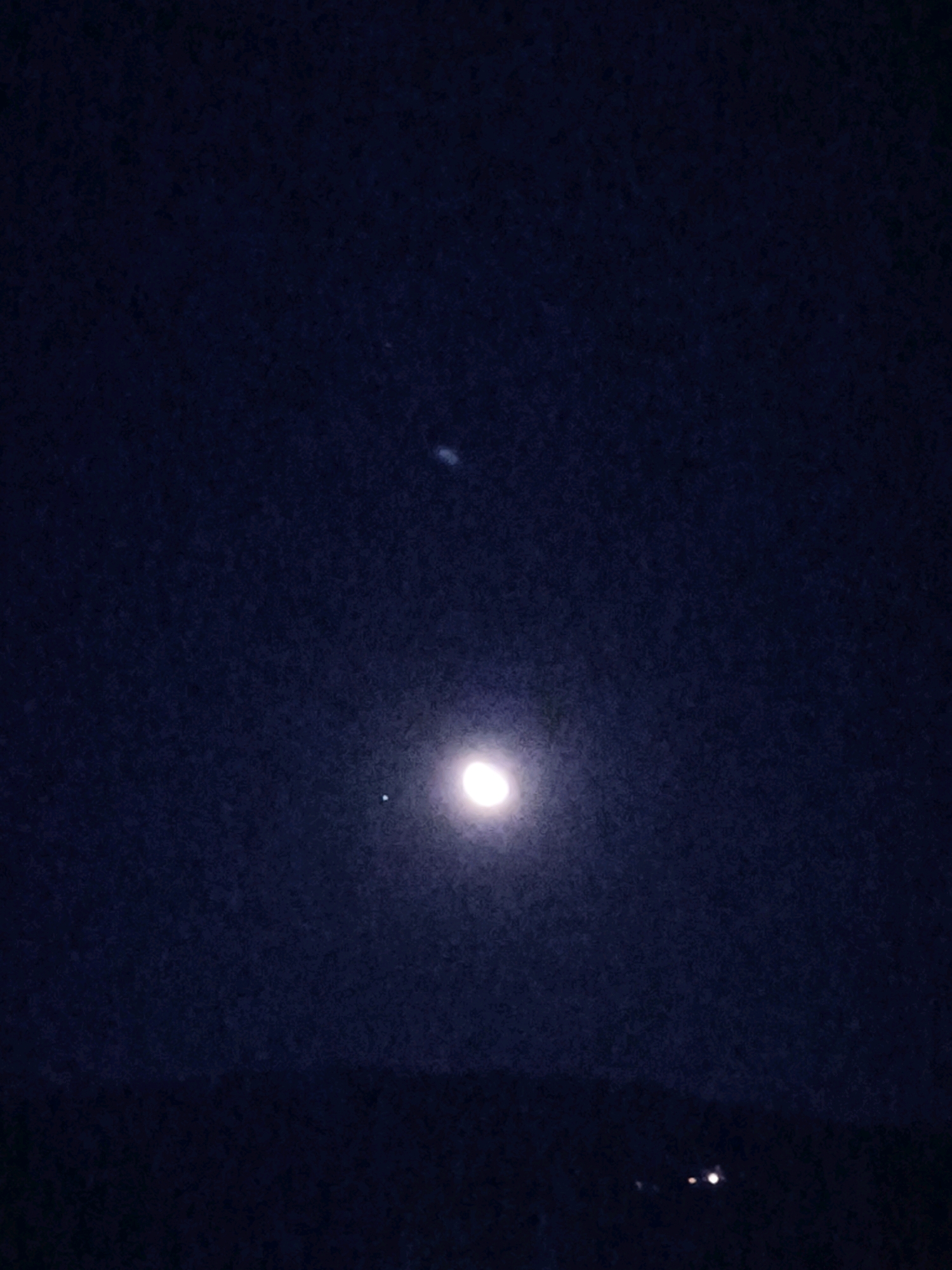 The image is a nighttime photograph of the sky above Passignano sul Trasimeno, Italy. The sky is dark blue and appears to be somewhat grainy. In the upper center of the image, there is a faint, elongated smudge of light, possibly a distant galaxy or cloud formation. Lower down, a bright, circular object, likely the Moon, is visible. To the left of the Moon, there is a smaller, distinct point of light, which could be a planet or a bright star. Near the bottom of the image, a dark, horizontal line suggests the silhouette of the distant landscape or horizon. On this horizon, there are two small, bright lights, possibly artificial lights from the town or vehicles. The overall impression is a nocturnal sky with celestial bodies and a hint of human presence.