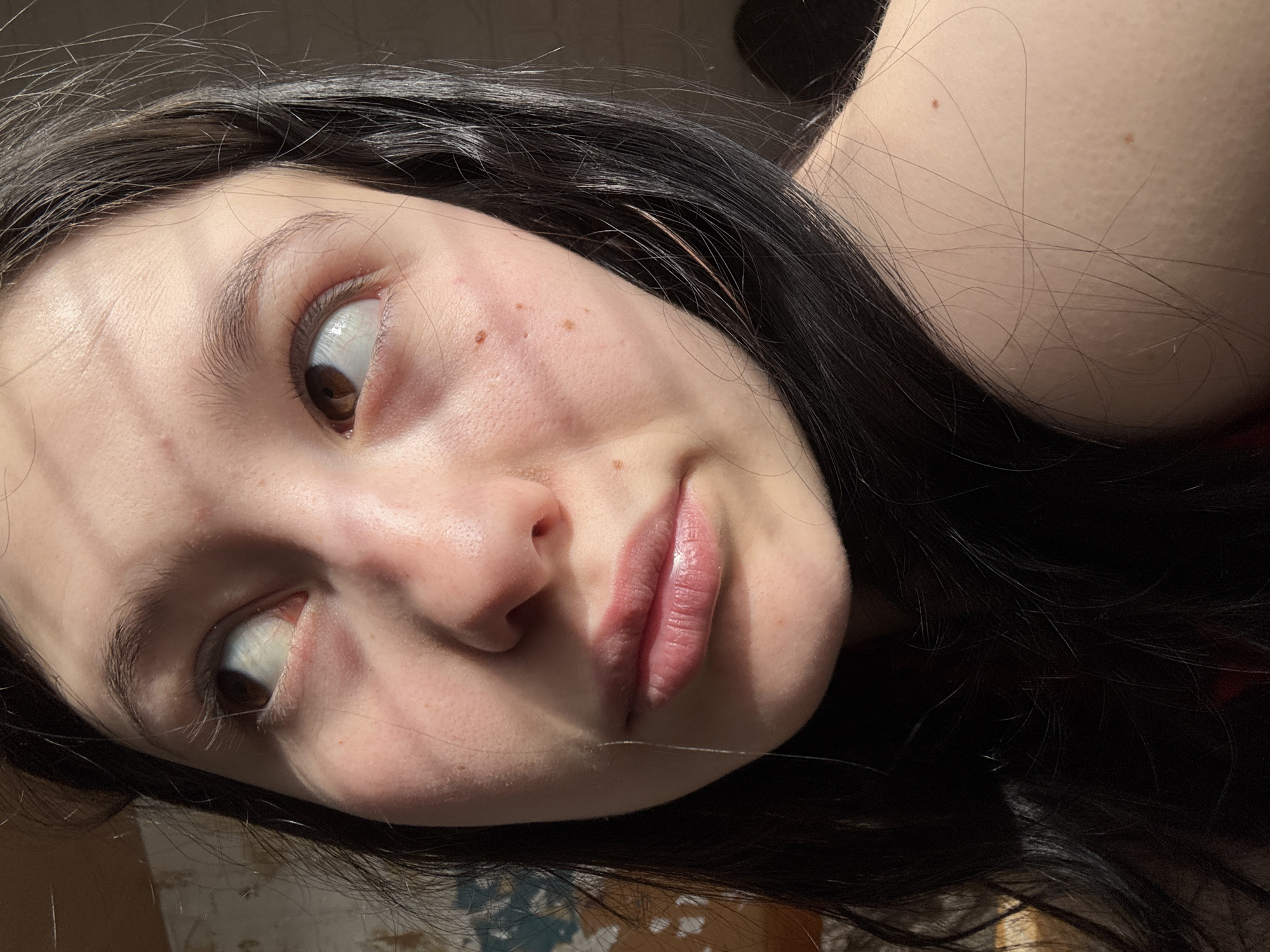 A close-up, angled portrait of a young woman with fair skin and dark hair. Her face is angled slightly towards the left, and her eyes are looking up and to the left. Her eyebrows are dark and thick. She has a small nose and full lips. The skin of her face has some freckles. There are shadows cast across her face, likely from blinds or a window frame. The background is indistinct, but there's a hint of a patterned surface near the bottom. It is not possible to determine the location of Фоки, Russia from this image.