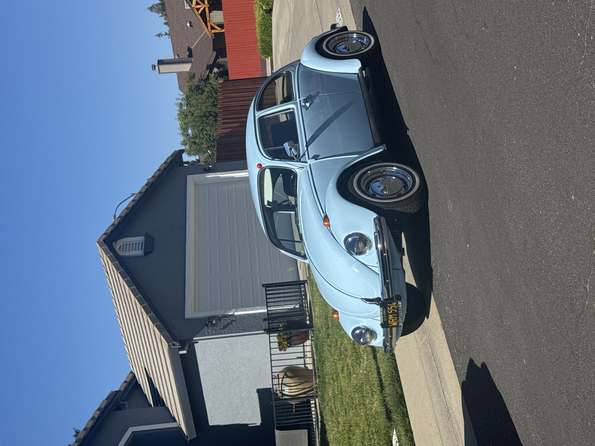 A light blue Volkswagen Beetle is parked on the right side of a paved street in front of a residential house. The car has chrome hubcaps and white-wall tires. The license plate reads "WRM 554". The house has a grey exterior and a white garage door. A black metal fence is in front of the house, with two decorative pots behind it. The street is bordered by a grassy lawn on the left. The sky is clear and blue. The image appears to have been taken in the city of Roseville, United States of America.