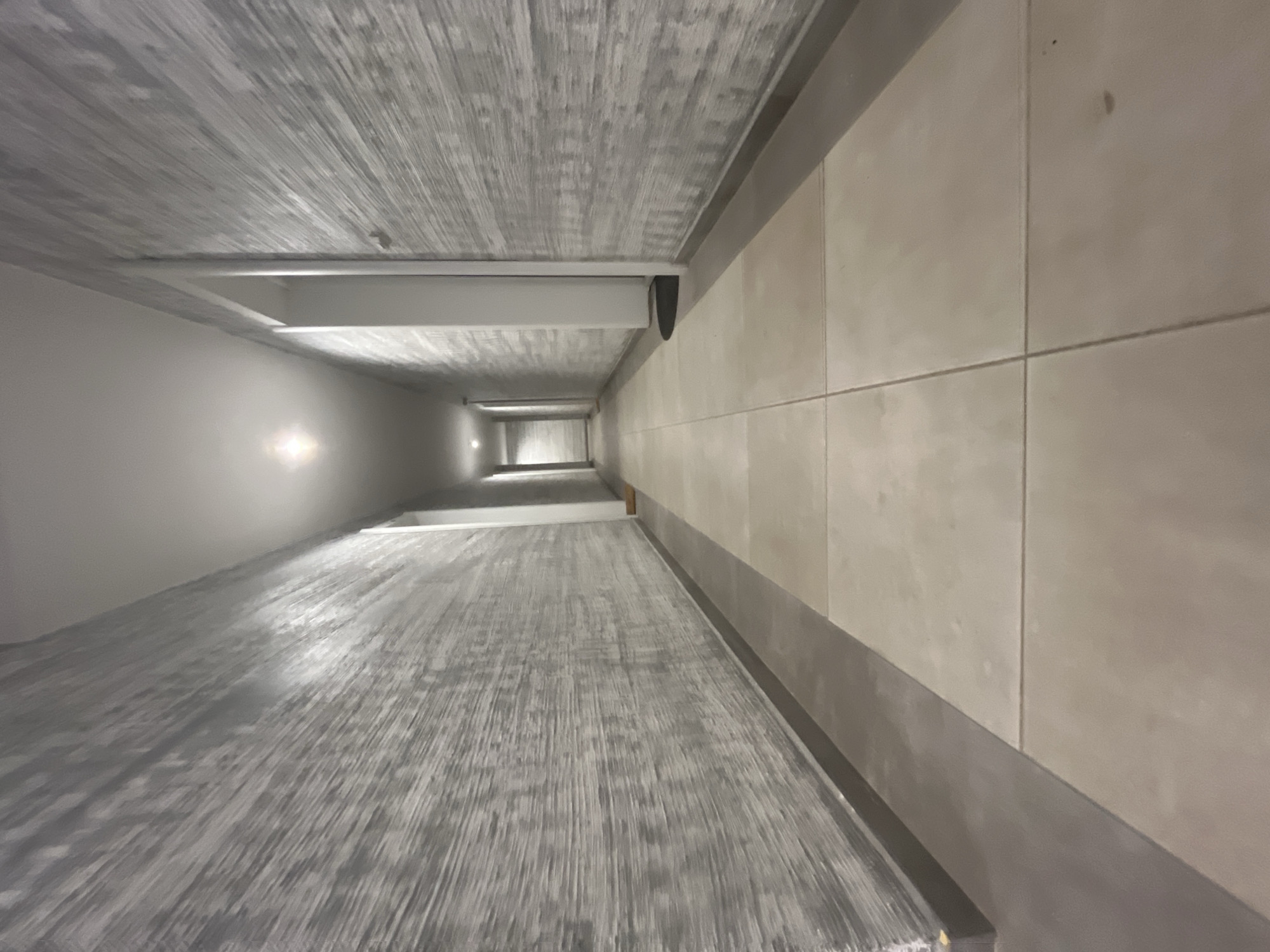 The image shows a long, narrow, and empty hallway with textured grey walls and light-colored floor tiles. The hallway extends into the distance, with a single light fixture illuminating the ceiling at its farthest visible point. There are doors visible on the right side of the hallway. The overall impression is one of a modern, minimalist interior. It is not possible to determine the specific location within the city of Varna, Bulgaria based solely on this image.