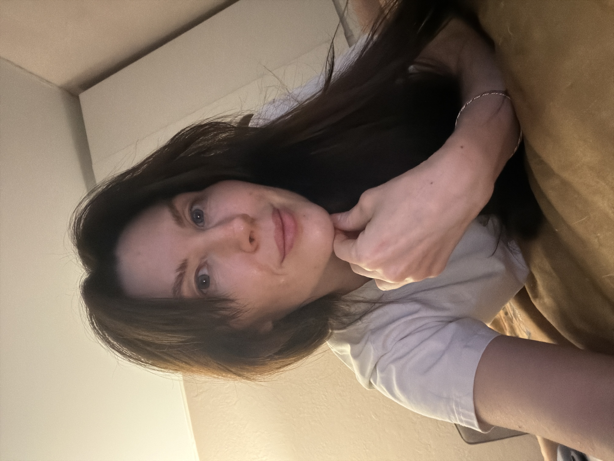 A young woman with long, dark hair and fair skin is lying on a brown surface, possibly a couch or bed, in a dimly lit room. She is wearing a light-colored t-shirt and a thin, silver bracelet on her right wrist. Her right hand is near her face, touching her chin. The walls are light-colored, and the room appears to be indoors. There is no evidence in the image to indicate it was taken in Perm, Russia.