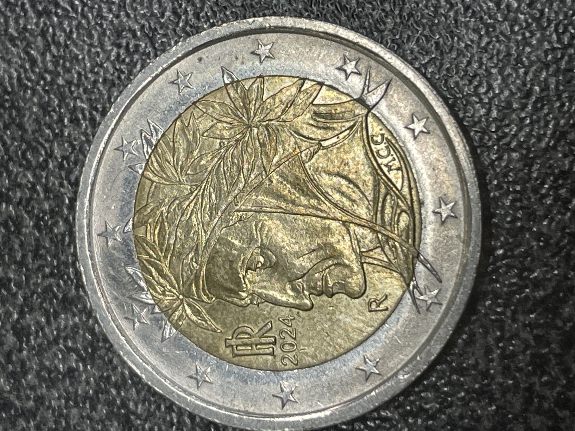 The image shows a close-up, high-angle view of a 2 Euro coin. The coin features a profile portrait of Dante Alighieri, the Italian poet, wearing a laurel wreath. The inscription "R" for Rome Mint appears on the lower left of the portrait, and the year "2024" is visible below the inscription "FR" (for Repubblica Italiana). The coin's edge is a silver color, and the central part is a gold color. The background is a dark, textured surface, possibly a table or cloth. There is no indication of Vienna, Austria, in the image.