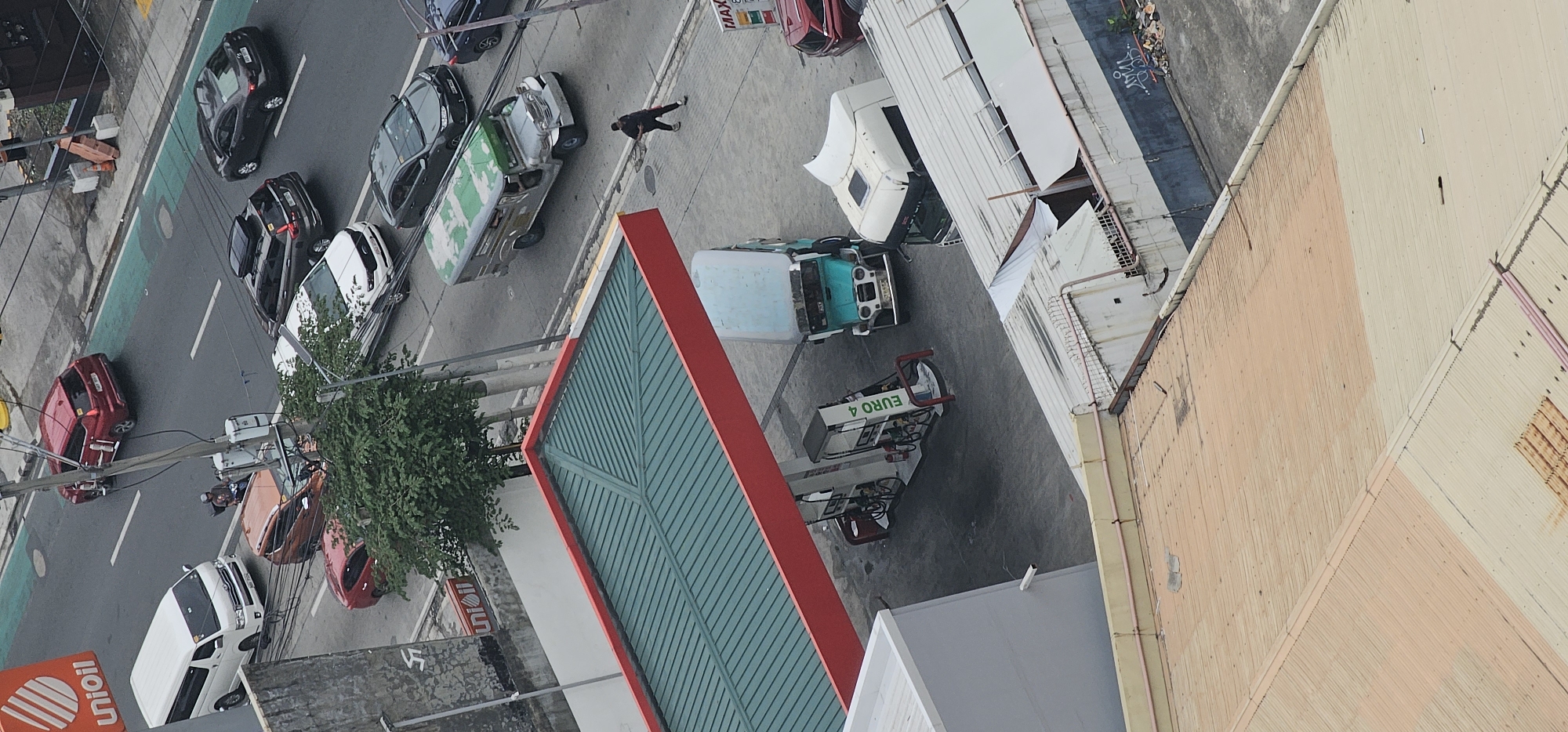The image captures an aerial view of a street scene in Makati City, Philippines, specifically near a UniOil gas station. The scene is characterized by a mix of vehicles, including cars, a van, a jeepney, and a truck, on a wet asphalt road. Several cars are parked or in transit, with one red SUV prominently visible on the road. A white van is parked near a building on the left. A jeepney, with a green roof, is positioned in the middle of the road, and a white truck is parked beside a gas pump.
A gas station with a green roof and red trim is visible in the foreground. Several gas pumps are situated under the canopy. A person in dark clothing is walking across the gas station's pavement, away from the road. Another jeepney, turquoise and silver, is parked near the gas pumps. In the background, more vehicles are seen on the road, including a dark gray sedan and a dark SUV with a roof rack. The street is lined with overhead wires and utility poles, typical of urban Philippine infrastructure. The general environment appears to be a busy urban setting with a mix of commercial and residential structures suggested by the buildings visible.