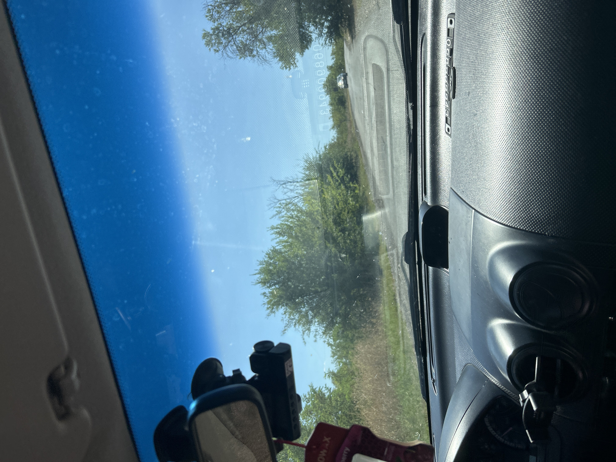 The image shows a view from the interior of a car, looking out through the windshield. The car appears to be driving on a paved road that curves to the right. The road is lined with trees and dry grass, suggesting a rural or suburban environment. A dark-colored car is visible in the distance, ahead and to the right on the road. The sky is bright blue with some scattered clouds, indicating daylight.
Inside the car, the dashboard is visible in the foreground. A dashcam is mounted on the windshield near the rearview mirror. A red air freshener is hanging from the rearview mirror. The interior appears to be a typical passenger vehicle.
The scene depicts a car journey on a sunny day, likely in or around Comrat, Moldova, given the context provided. There are no people clearly visible in the image. The focus is on the road ahead and the surrounding natural environment.