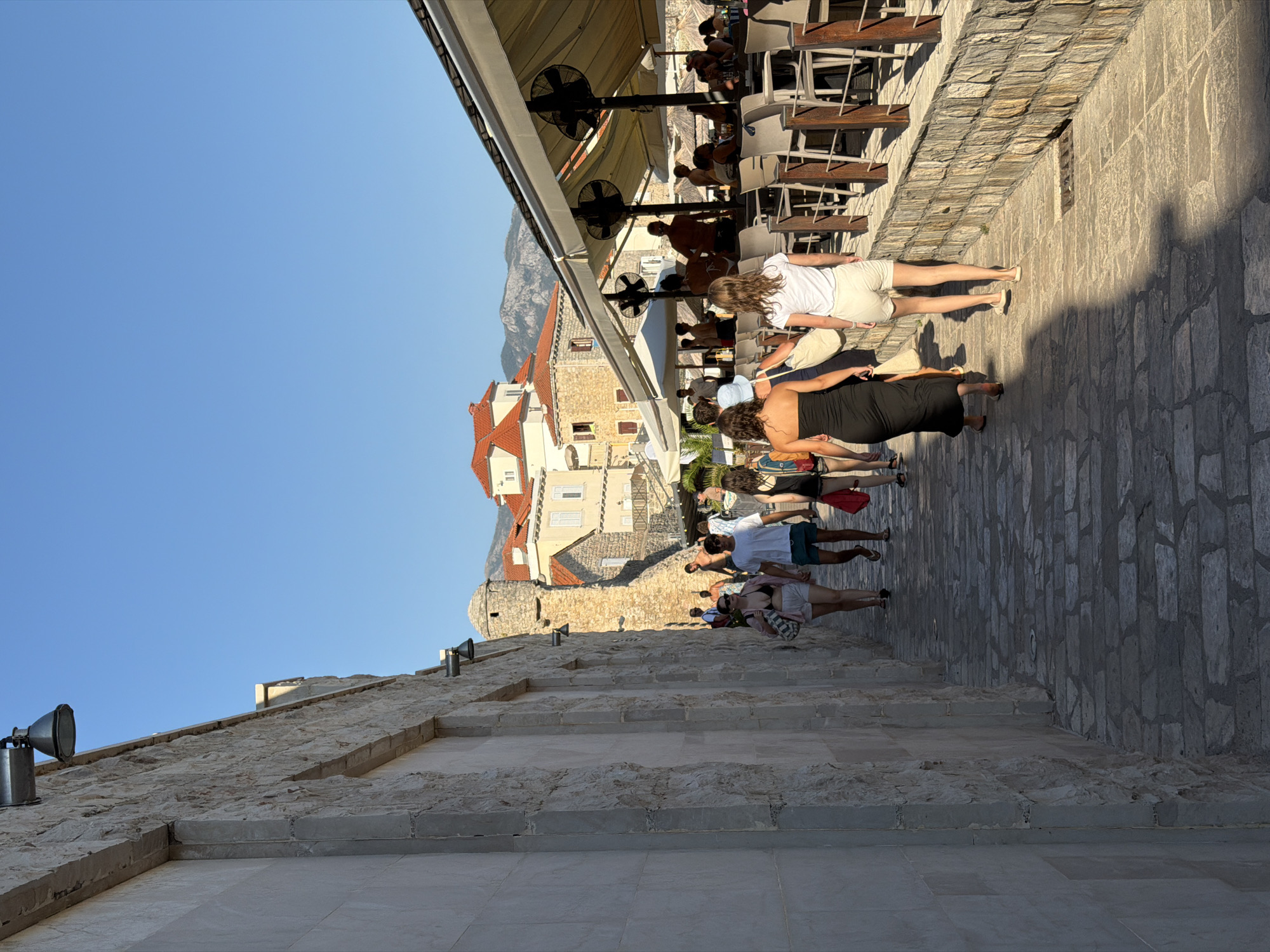 The image captures a sunlit street scene in the old town of Budva, Montenegro, likely along the promenade. A group of people are walking down a cobblestone path, casting long shadows. The architecture is characteristic of the region, with stone buildings featuring terracotta roofs visible in the background. On the right side of the path, there is an outdoor seating area for a restaurant or cafe, with tables, chairs, and large umbrellas providing shade. The atmosphere appears to be relaxed, with individuals dressed in casual summer attire, suggesting a tourist destination during warm weather. The stone wall on the left side of the image is a prominent feature, suggesting a fortified area or an older structure.