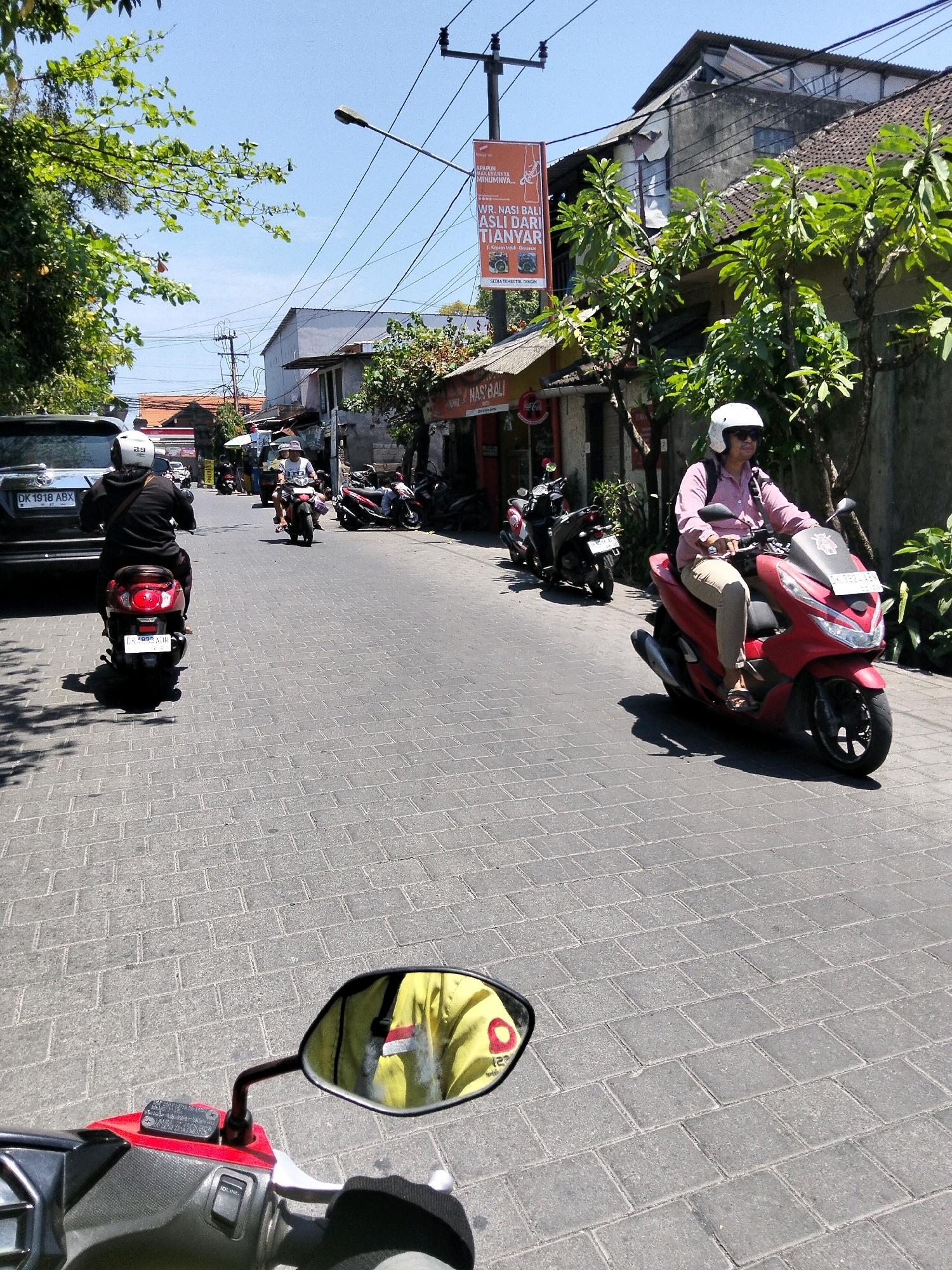 The image depicts a street scene in Denpasar, Indonesia, likely on a sunny day. In the foreground, the handlebars and a portion of the body of a red scooter are visible, with a side mirror reflecting the rider's yellow jacket, which has an Indonesian flag emblem. The rider is wearing a white helmet and sunglasses. The scooter is on a paved road made of interlocking gray bricks.
Further down the street, several other motorcycles are present. A red scooter is being ridden by a person wearing a pink shirt and a white helmet. Behind them, a black scooter is also visible. To the left, a black car with a license plate starting with "DK" is parked, and a person wearing a black hoodie and a white helmet is riding a red scooter away from the car.
On the right side of the street, there are buildings with shops and trees. A prominent banner advertising "NASI BALI ASLI DARI TIANYAR" is displayed on a pole. The buildings appear to be a mix of traditional and more modern structures. Electrical wires crisscross the sky, typical of urban Indonesian environments. The overall impression is a typical day of street life in a bustling Indonesian city.