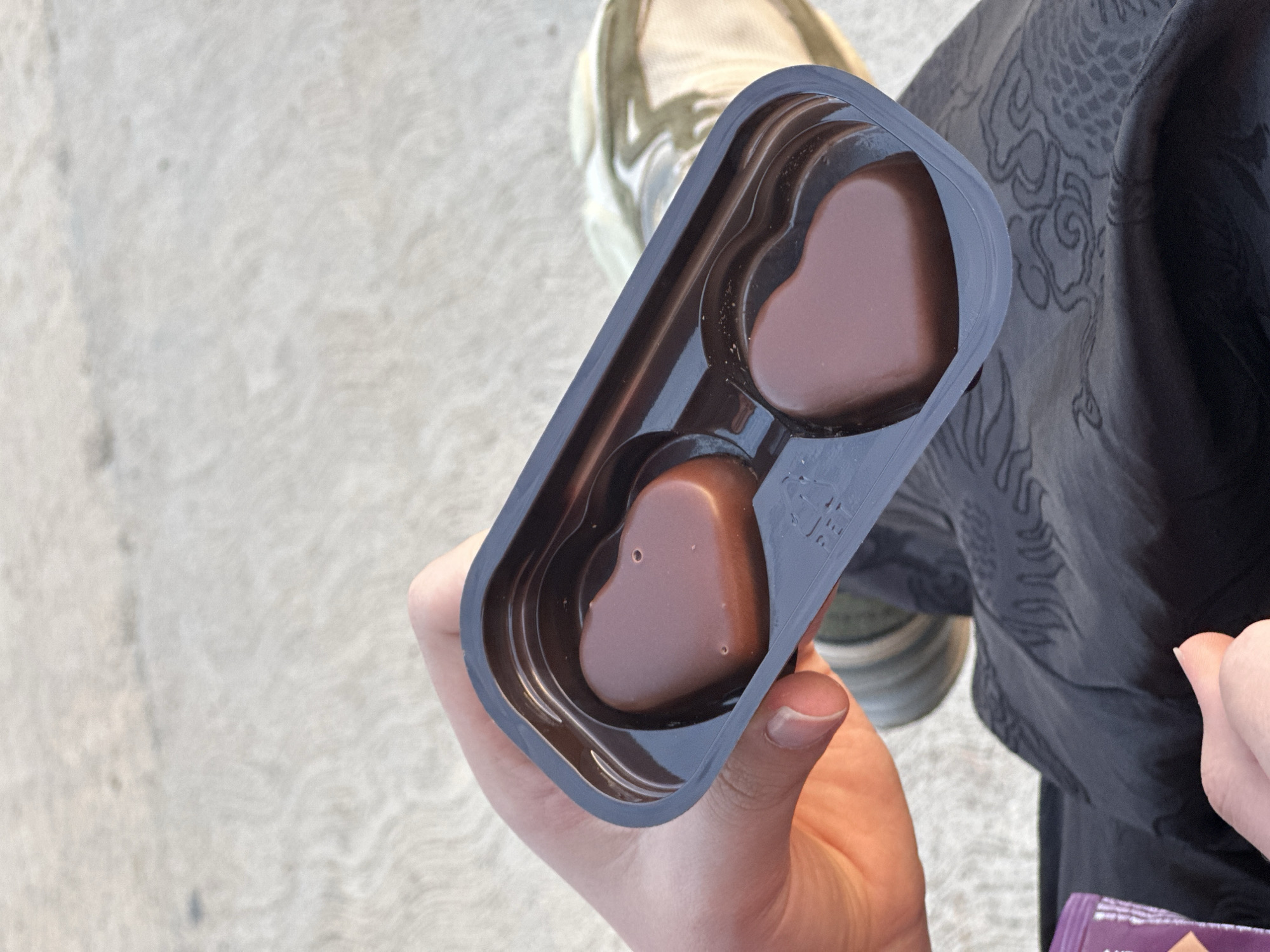 An overhead, slightly angled shot shows a person's hand holding an open plastic container with two heart-shaped milk chocolates. The container is dark brown or black plastic, designed to hold the chocolates snugly. One chocolate, closer to the viewer, has small indentations or air bubbles on its surface. The top part of the container is being peeled back to reveal the candies.
The hand holding the container is light-skinned, with visible knuckles and short, clean fingernails. In the background, mostly out of focus, is light grey, textured ground, likely concrete or pavement. To the top left, part of a light-colored shoe or sneaker is visible. To the right, a dark piece of clothing, possibly trousers or a skirt, is visible with an intricate, lighter grey patterned design, resembling swirling clouds or a dragon. In the very bottom right corner, a sliver of a purple wrapper with white text is partially visible, too blurry to read fully, but appears to contain the letters "AN" and possibly "U" along with a small symbol. The scene takes place outdoors during daylight hours in Šušanj, Montenegro.