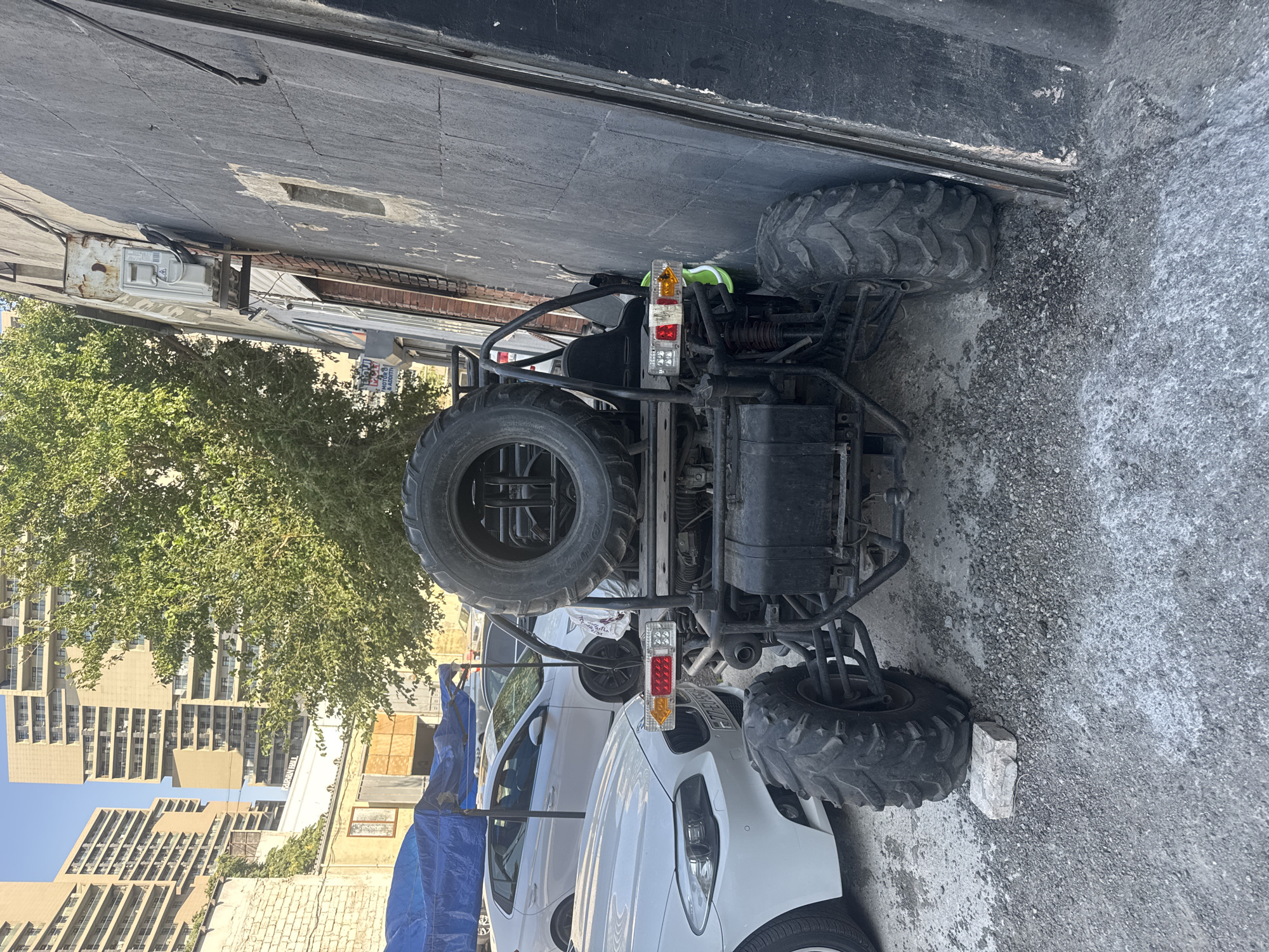 An all-terrain vehicle (ATV) is parked on a gravel surface next to a dark gray building. The ATV is black and has a spare tire mounted on its rear. It also features prominent suspension and large, knobby tires. In the background, a white sedan is parked. Further back, a blue tarp covers some equipment, and behind that, several modern, multi-story buildings are visible under a clear sky. The scene is likely in Baku, Azerbaijan, given the architectural style of the buildings. There are no people in the image.