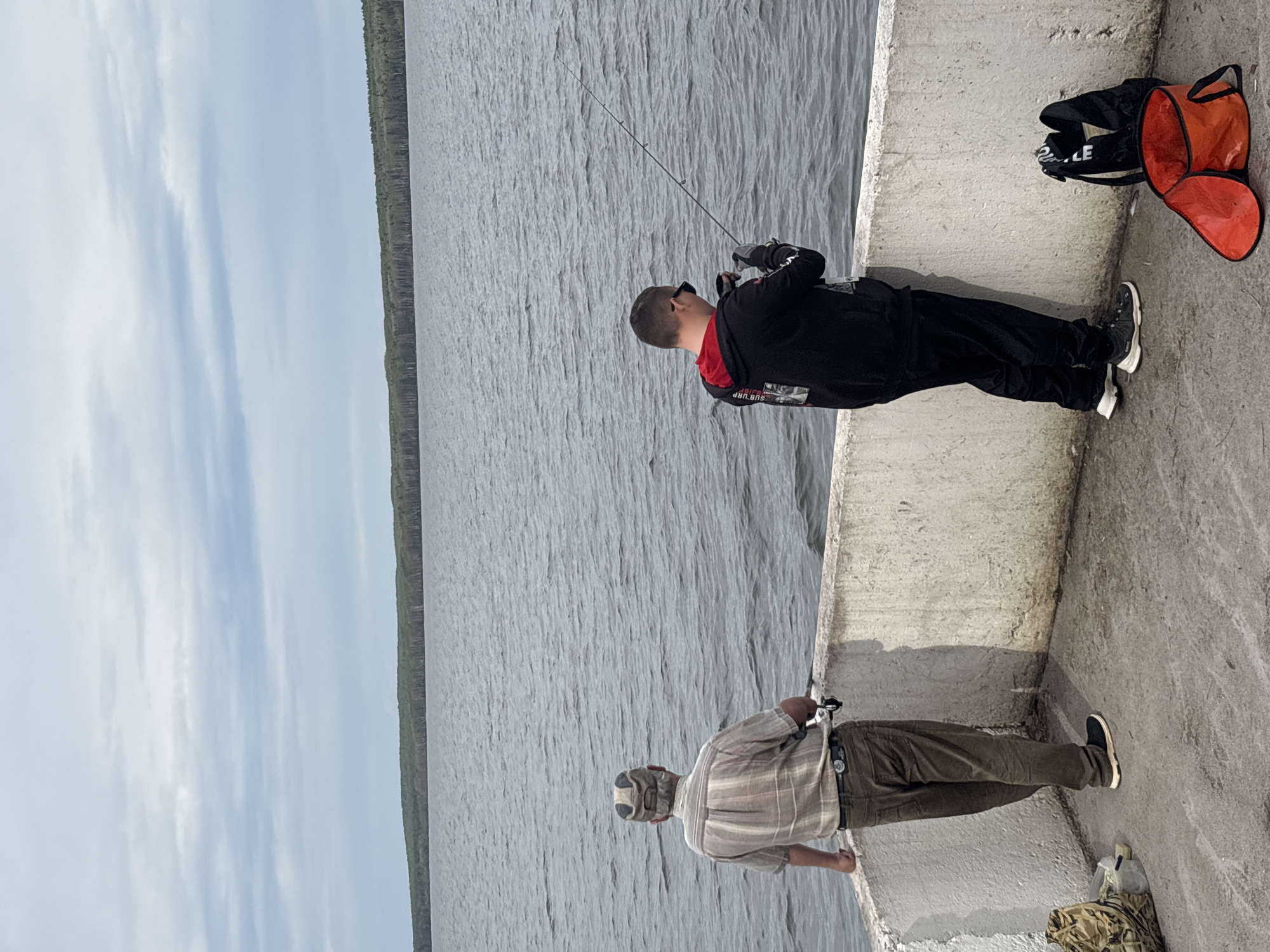 Two men are fishing on a concrete pier. The pier extends into a large body of water, likely a lake or reservoir, with a forested shoreline visible in the distance. The sky is overcast. One man, closer to the camera, wears a plaid shirt, khaki pants, and a baseball cap. The second man, further away, is wearing a black jacket with red accents, black pants, and sunglasses. Both men are holding fishing rods. An orange and black bag sits on the pier near the second man. The location is likely near Chaikovsky, Russia, given the request.