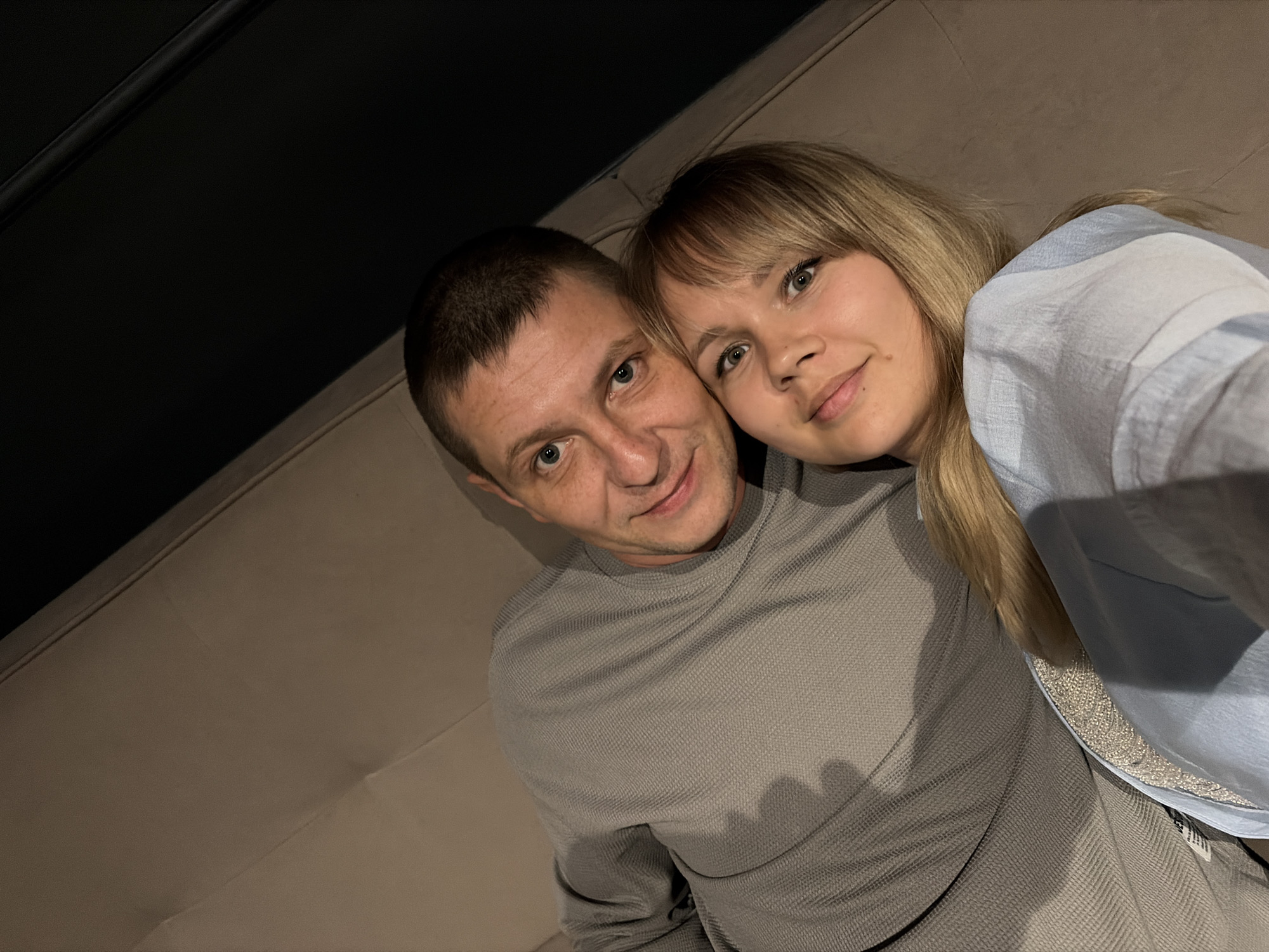 In Chaikovsky, Russia, an overhead indoor photograph depicts an adult male and an adult female reclining on a light beige upholstered surface. The male, with short dark hair and light eyes, is positioned on the left side of the frame, wearing a grey textured long-sleeved top, and has a slight smile. The female, with blonde hair and bangs and light eyes, is positioned in the foreground and to the right, wearing a light blue long-sleeved shirt over an inner patterned layer, also displaying a slight smile. The woman's head rests near the man's. Both individuals are looking directly at the camera. The upper right portion of the background is dark, featuring a thin dark horizontal line, possibly a structural element or trim.