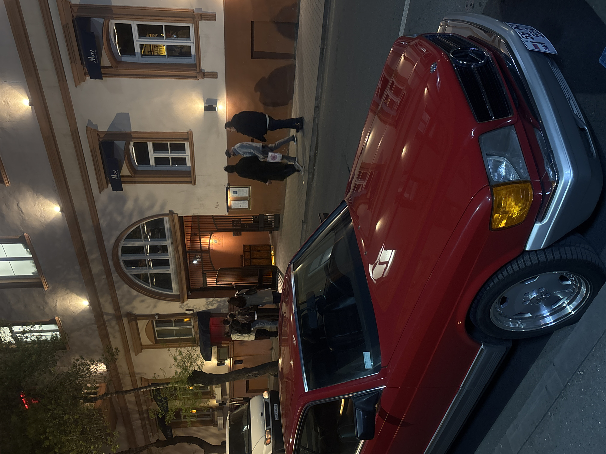The image shows a street scene in Yerevan, Armenia at night. In the foreground is a red vintage Mercedes-Benz parked on the right side of the road. Behind the car, several people are entering a building, possibly a restaurant or bar, as indicated by the awning with "Mov" written on it. The building's facade has ornate window frames and architectural details illuminated by streetlights. A white van is parked along the street behind the Mercedes. People are walking on the sidewalk past the building.