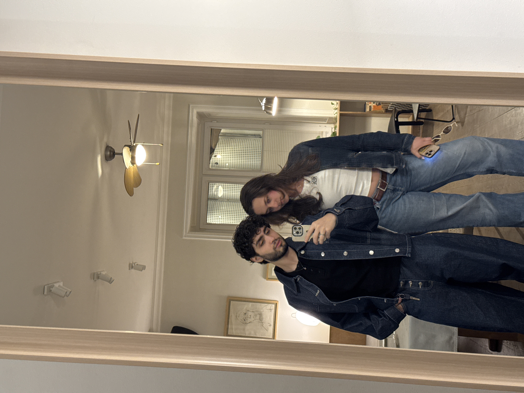 The image is a reflection of a man and woman posing for a photo in what appears to be an apartment or house. The man, who has dark curly hair and a beard, is holding up a phone to take a picture of the couple. He is wearing a black shirt and a dark denim jacket and pants. The woman, who has long brown hair, is leaning her head on his shoulder. She is wearing a white top and blue jeans with a brown belt, holding sunglasses in her left hand. The reflection reveals details of the room, including a light fixture with a fan, wall-mounted lighting fixtures, a framed piece of art, a window, and furniture in the background. There is no evidence to suggest this photo was taken in Yerevan, Armenia.