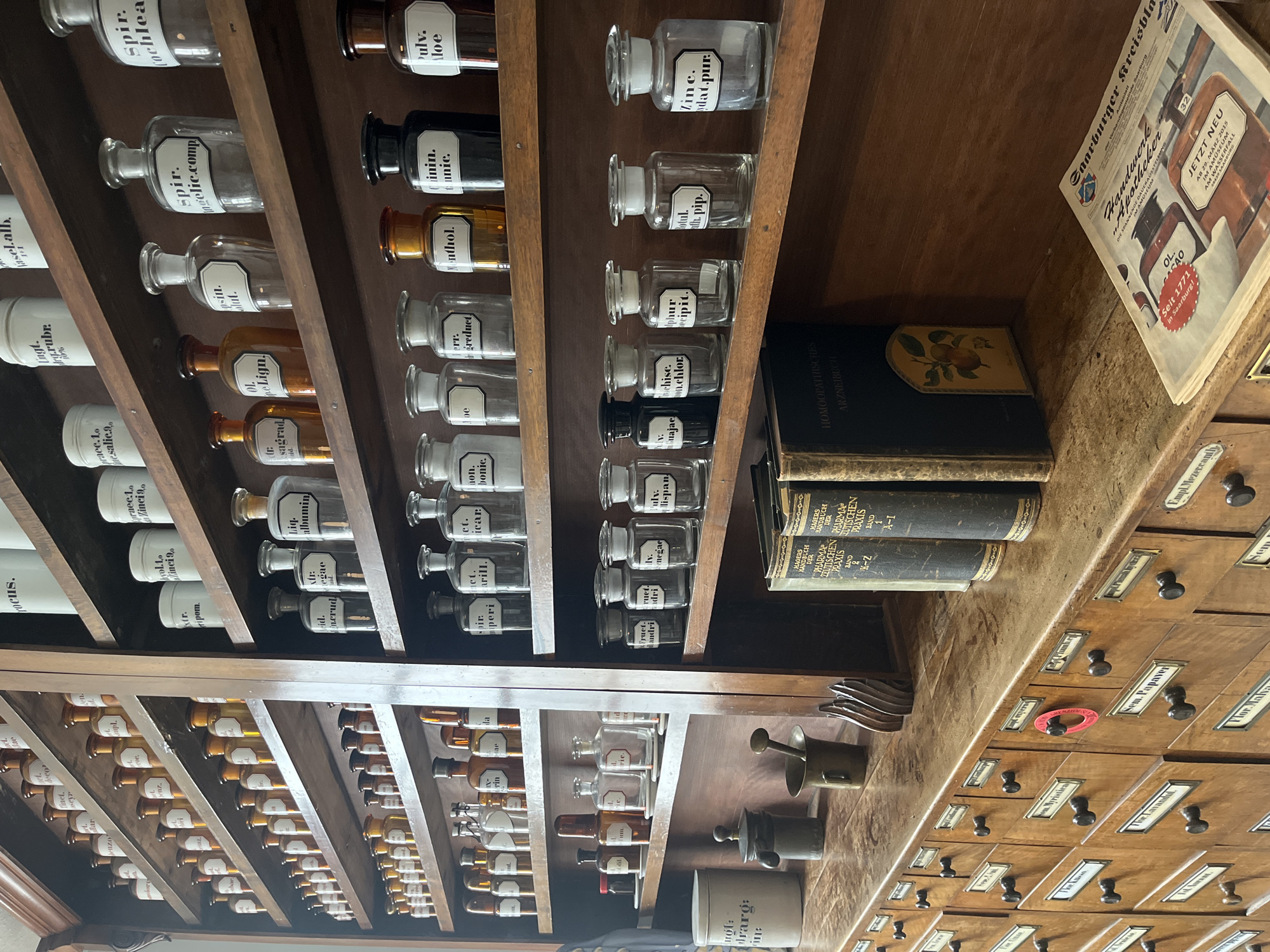 The image depicts a vintage apothecary setup, likely within a museum in Saarburg, Germany. Shelves are filled with numerous labeled glass bottles and jars containing various medicinal ingredients. Below these, wooden drawers with labeled compartments suggest an organized storage system for further supplies. Books related to homeopathy and pharmaceutical practices lie adjacent to the apothecary items. A newspaper advertises a permanent exhibition about the history of apothecaries in Saarburg.