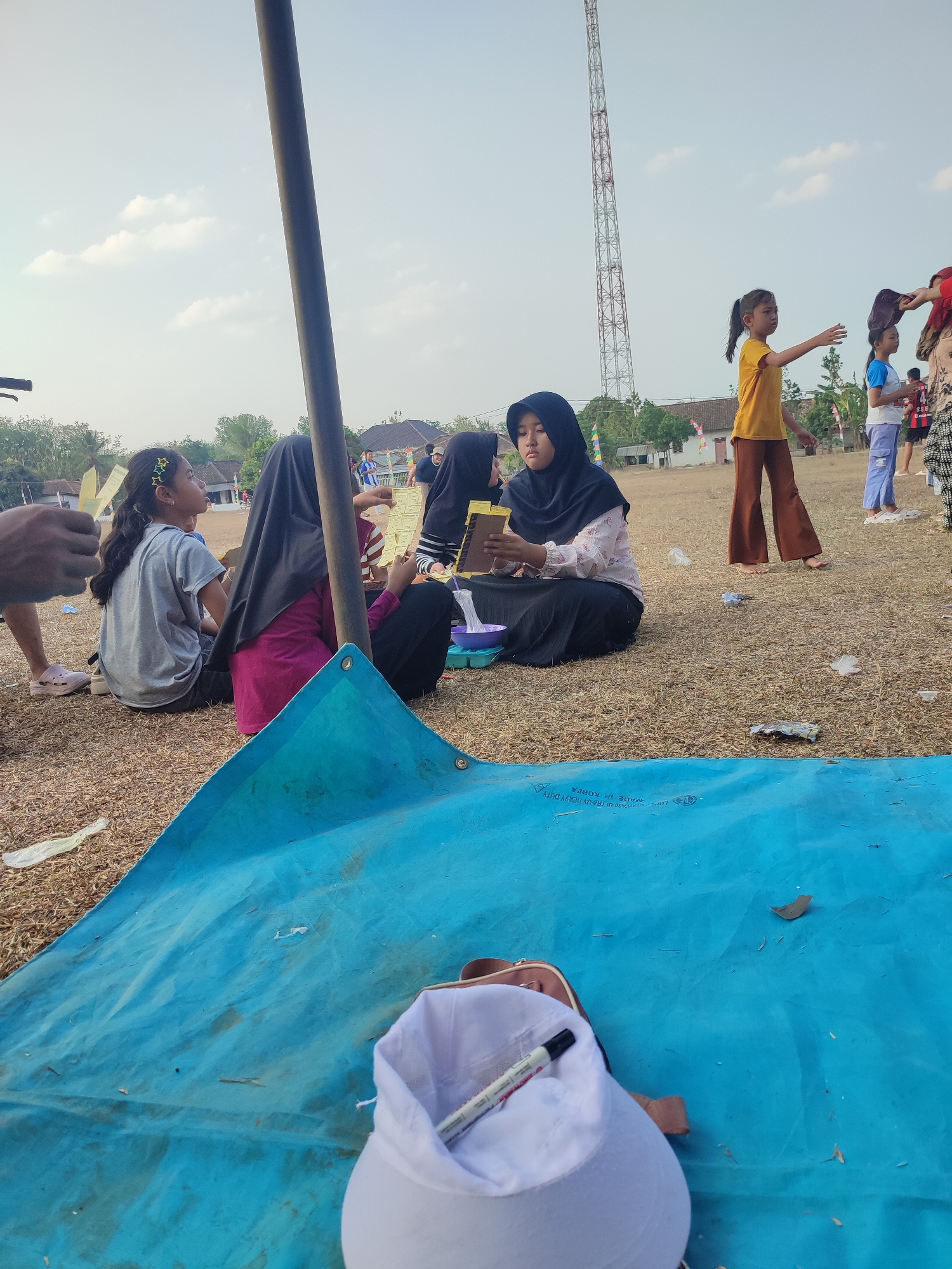 In Nusa Jaya, Indonesia, on a dry, dusty field, several individuals, predominantly young girls, are gathered outdoors. A tall, lattice-like tower pierces the hazy sky, dominating the background. In the foreground, a large, bright blue tarp lies spread out on the ground, partially obscuring a white sun hat with a marker pen. Several children are seated on the dry grass; one girl in a gray t-shirt and another in a black hijab are looking at small objects. Further back, a girl in a yellow shirt and brown pants stands, interacting with another child in a blue shirt. Scattered debris, including plastic wrappers and leaves, litter the ground. The overall impression is of a casual outdoor gathering or event.