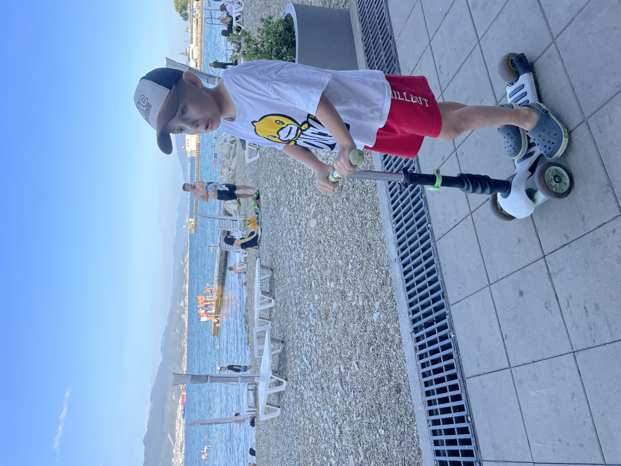 A young boy wearing a white t-shirt with a yellow duck graphic, red shorts, a baseball cap, and grey Crocs rides a three-wheeled scooter on a paved walkway. He is positioned in the foreground, with a beach and the sea in the background. The beach is covered in pebbles, with several white lounge chairs and umbrellas. Several people are visible on the beach and in the water. The sea is calm, with mountains in the distance. The sky is blue with a few scattered clouds. The image appears to have been taken in Ivanovskaya, Russia, based on the provided location context.