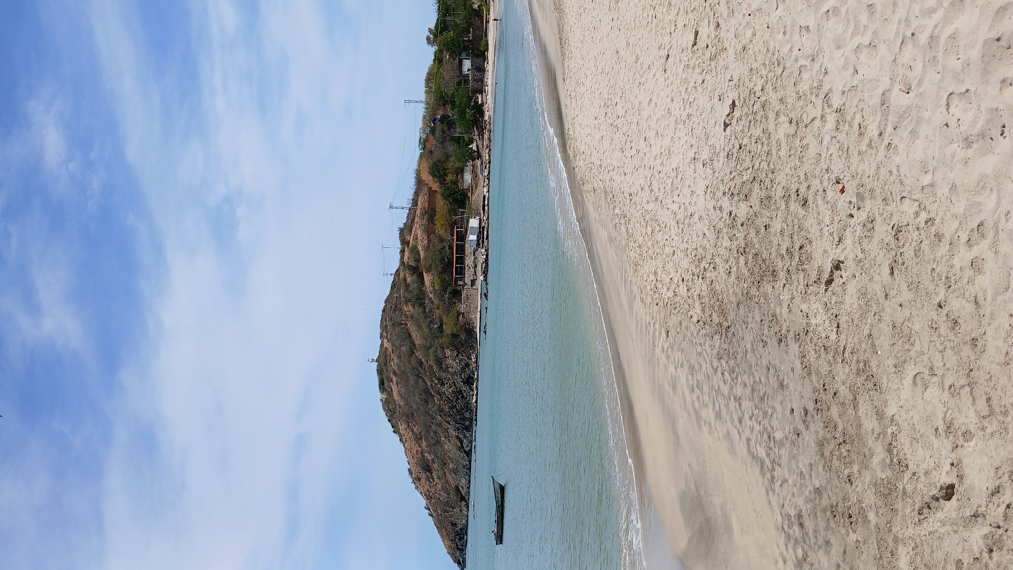 The image displays a coastal scene in Pedro González, Venezuela. The foreground consists of a wide, light tan sandy beach with visible footprints and minor textural variations. Gentle, light blue-green ocean water with small waves laps the shore along a curved waterline. A dark-colored, small boat is anchored approximately one-third from the left edge of the frame, floating in the calm water.
In the background, a prominent, arid-looking hill covered with sparse brown and green vegetation rises. A red and white striped lighthouse or tower is situated on the hill's peak, slightly left of the central axis. Below the peak, power lines with multiple poles traverse the hillside. At the base of the hill, closer to the right side of the frame, several small structures with white walls and reddish-brown roofs are visible, indicating a developed area near the shoreline. A single, small human figure is discernible walking along the distant right edge of the beach where it meets the water. The sky above is light blue with scattered white clouds.